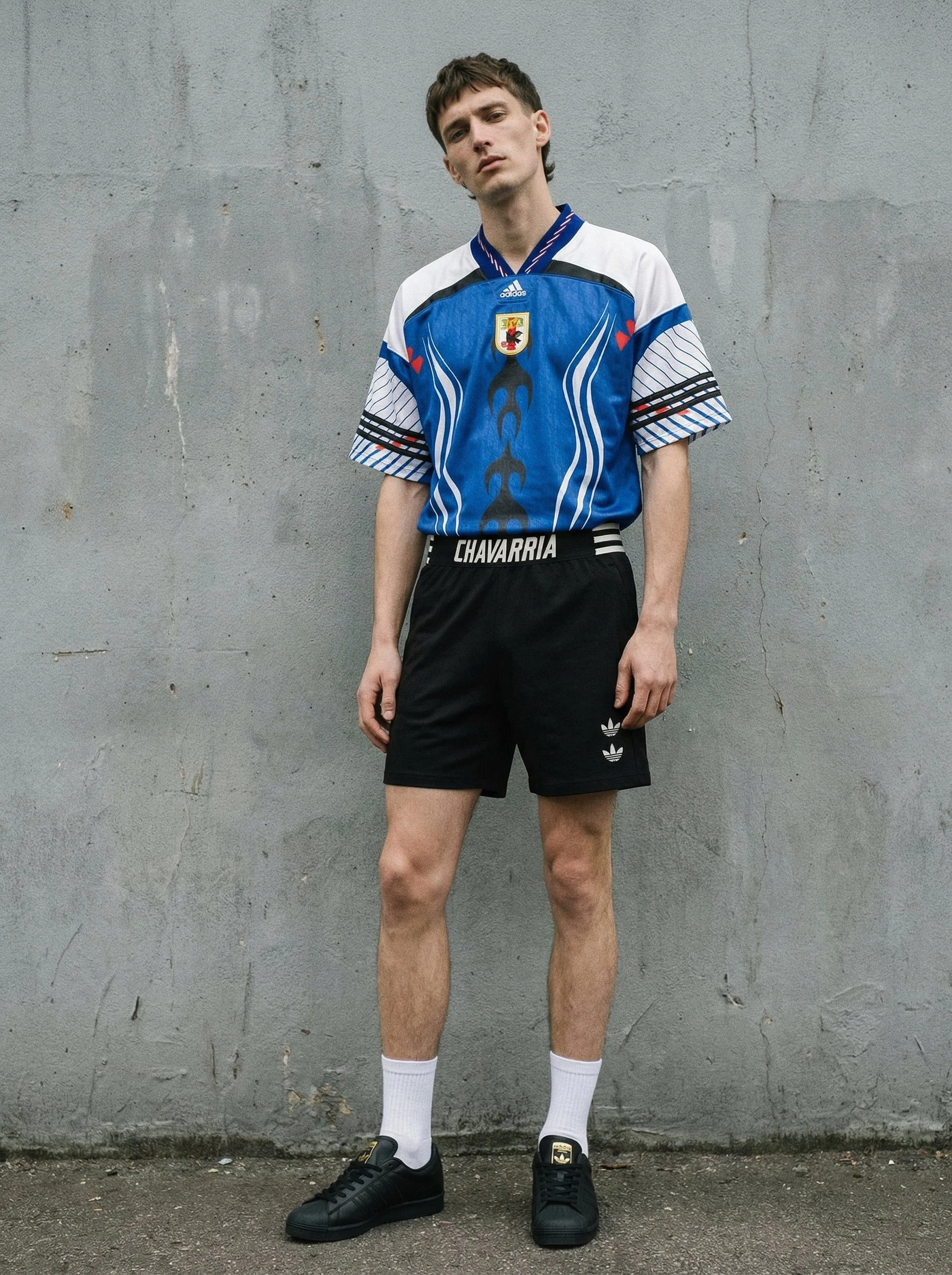 A young man with short brown hair standing against a plain gray concrete wall, wearing a blue, white, and black sports jersey, black shorts, white socks, and black Adidas sneakers.
