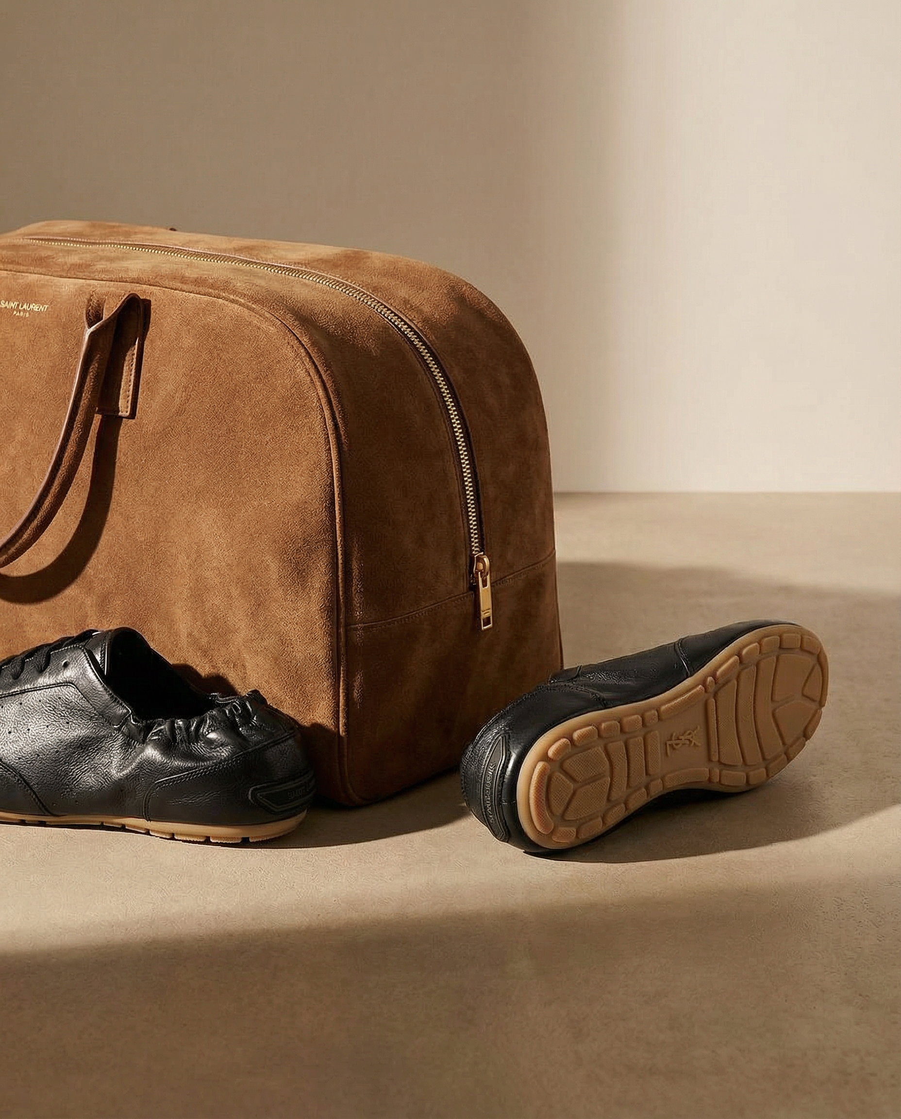 A brown leather of Saint Laurent Paris suitcase with a zipper and handles, placed on a flat surface, with a pair of black sneakers with beige soles lying next to it.