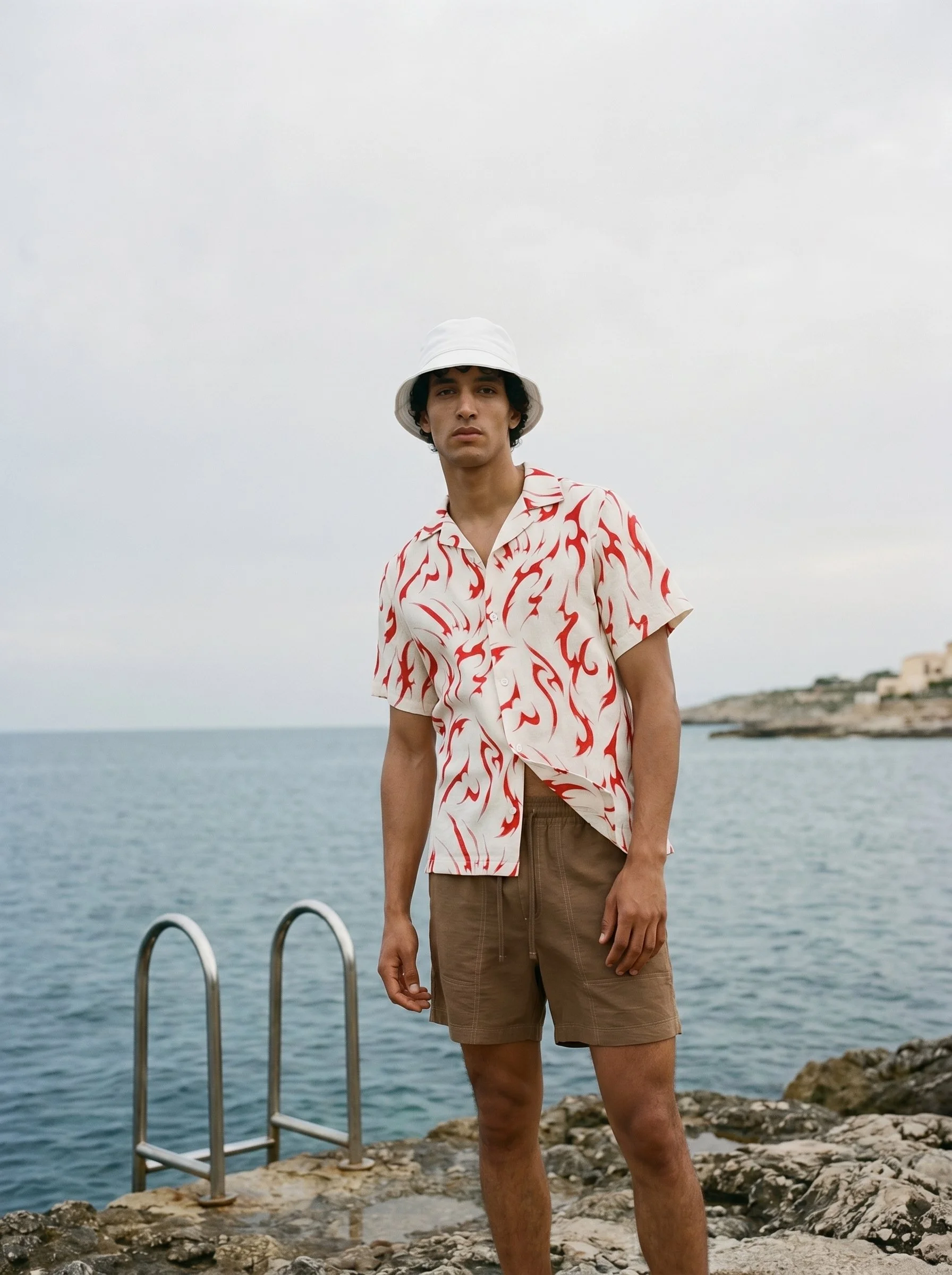 A young man standing by a rocky shoreline near water with a metal ladder, wearing a white bucket hat, a red and white patterned short-sleeve shirt, and brown shorts, with a hilly coastline in the background.
