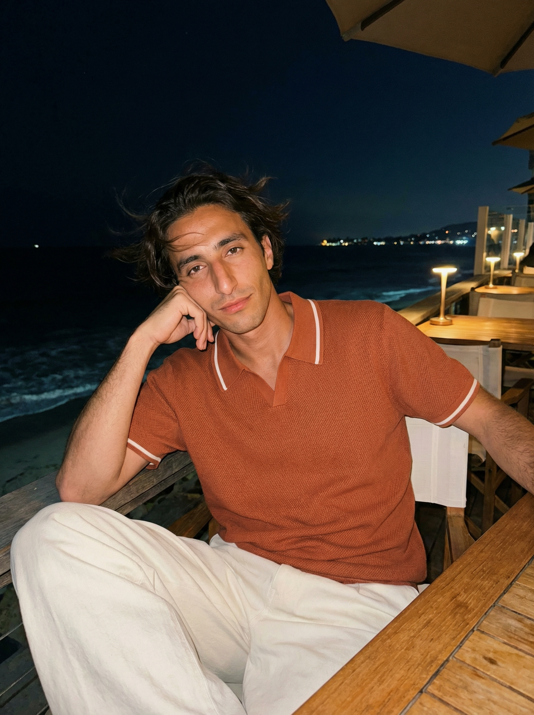 A man sitting at an outdoor dining area at night, with ocean and distant city lights in the background, wearing an orange shirt and white pants, resting his head on his hand.
