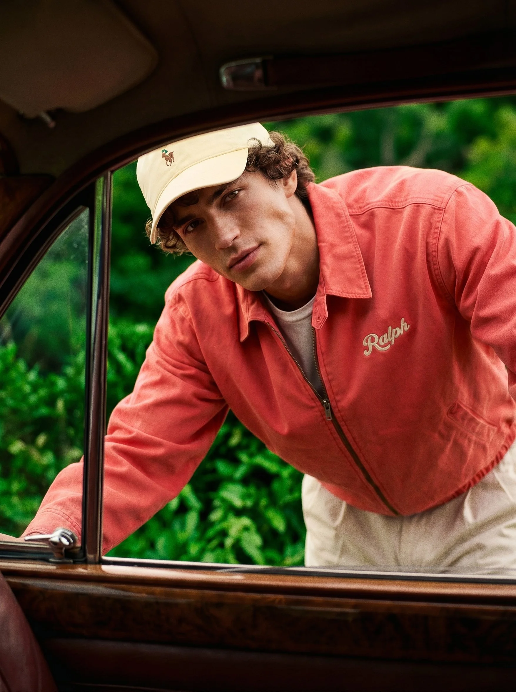 A young man with curly hair wearing a beige cap and coral jacket, leaning into a car window with a green, leafy background.