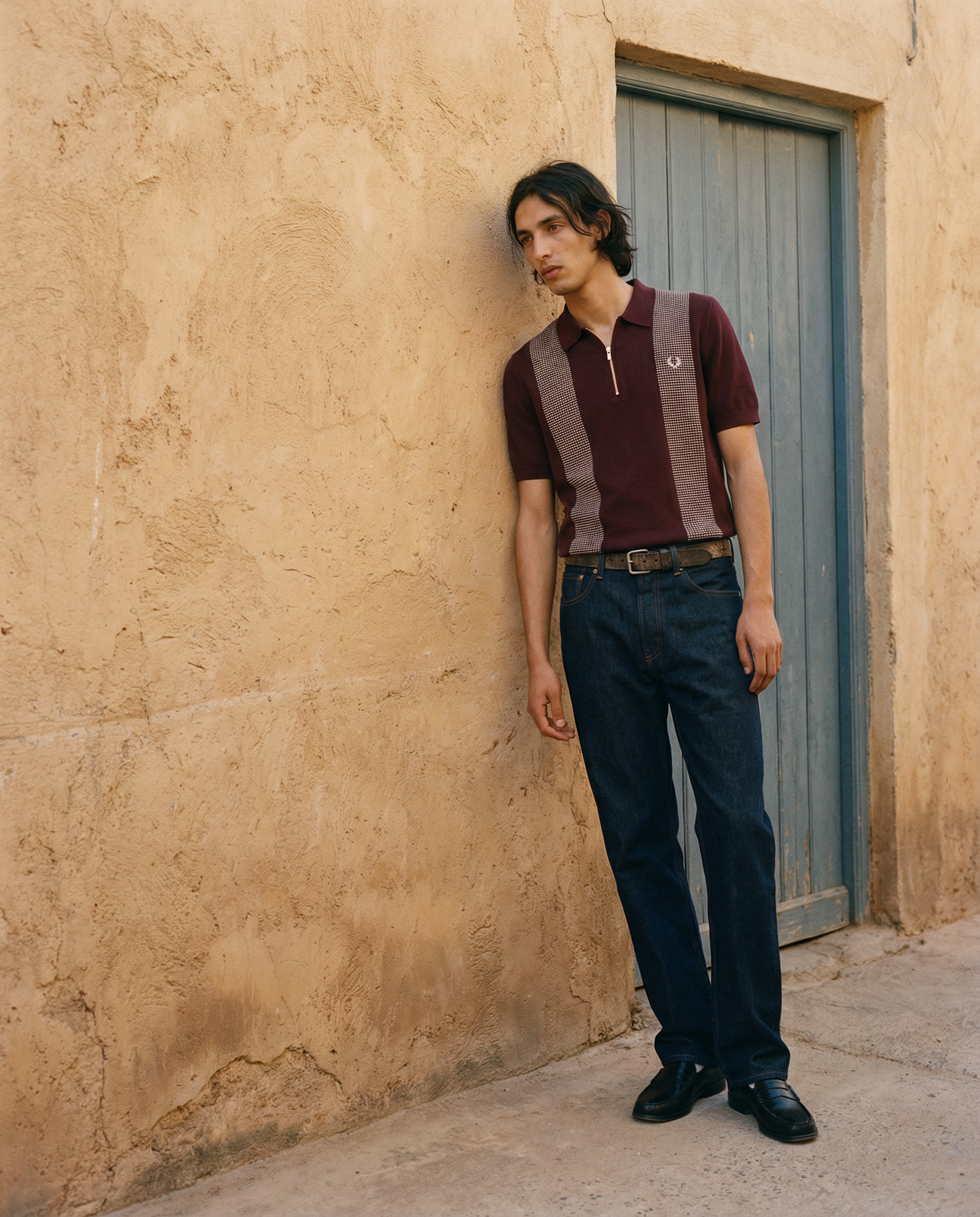 A young man with long dark hair leaning against a textured beige wall with a closed blue door behind him, wearing a maroon shirt with a half zipper and dark blue jeans.
