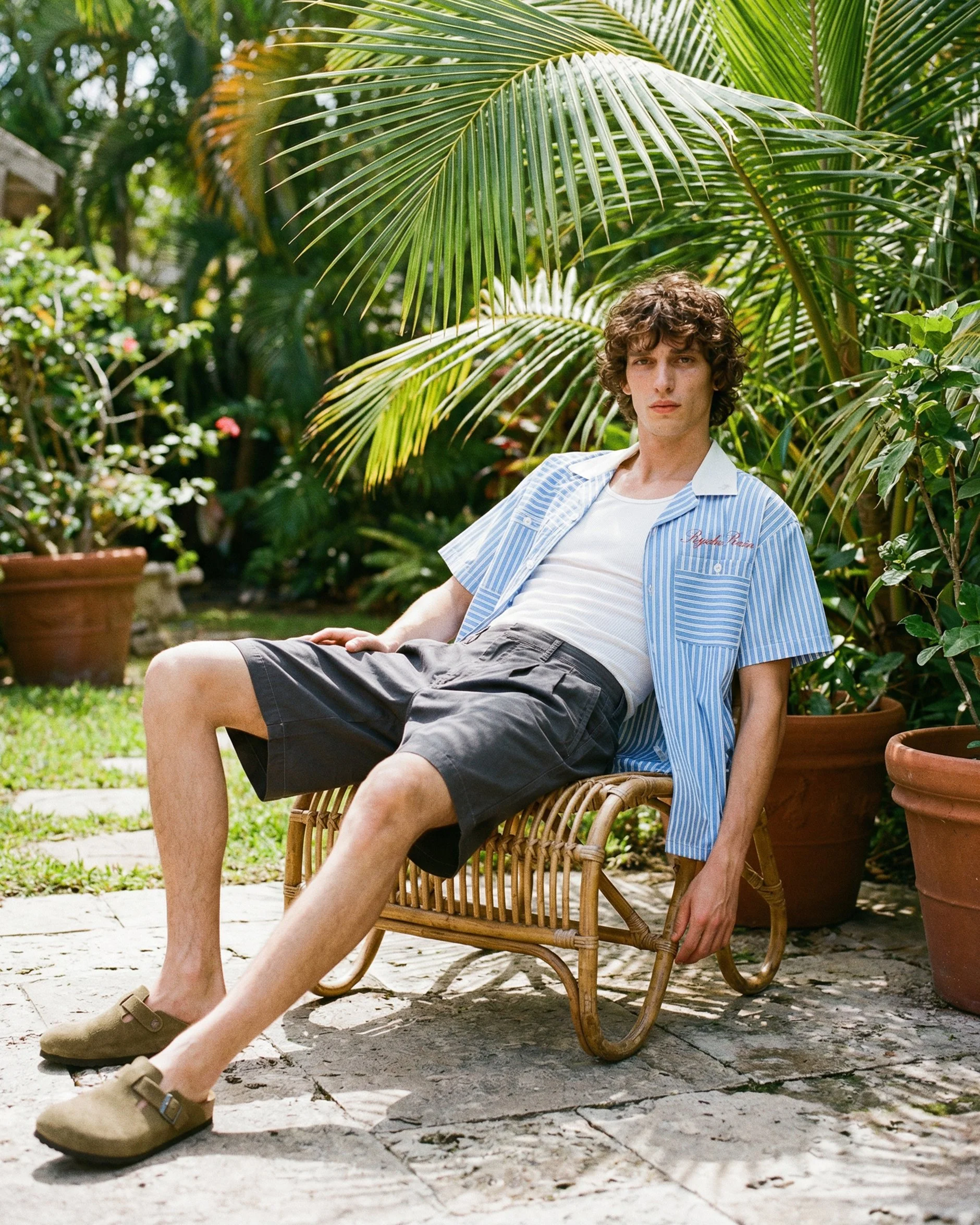 A young man with curly hair is sitting on a wicker chair outdoors, surrounded by large green potted plants and palm leaves, wearing a light blue, short-sleeved, striped shirt, white T-shirt, black shorts, and tan slip-on shoes.  AI fashion editorial 