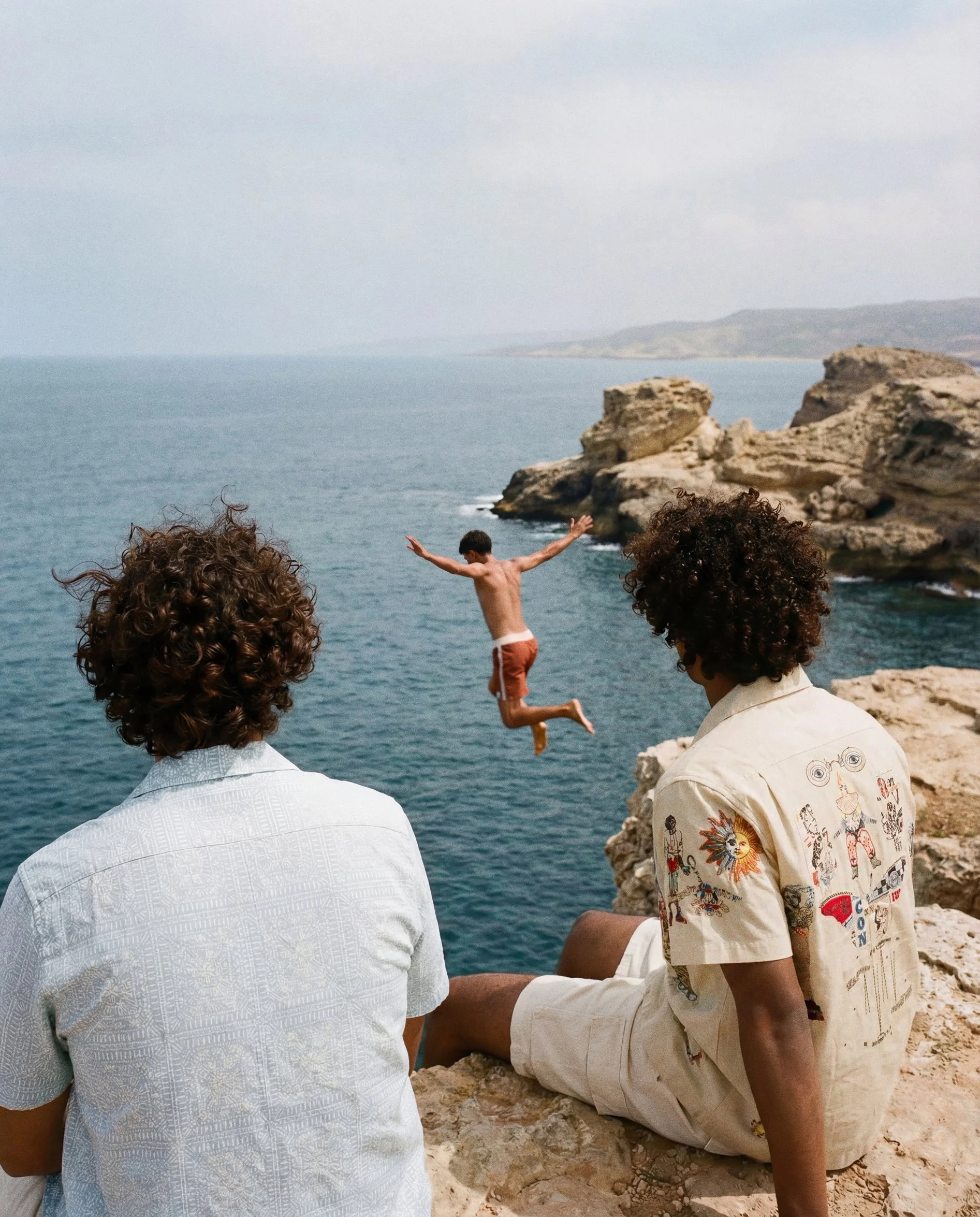 People sitting on rocks by the ocean, one person jumping into the water, with rocky cliffs and a cloudy sky in the background.