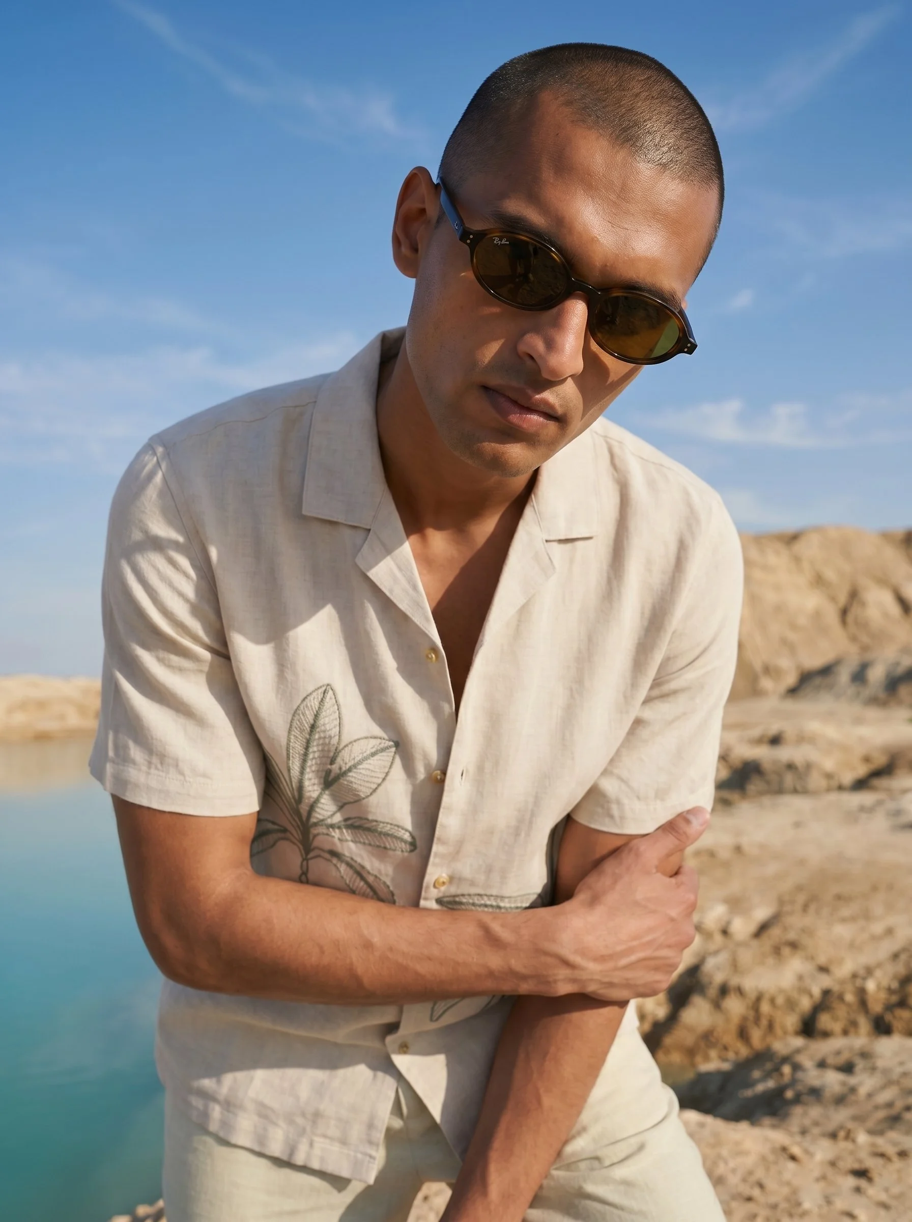 A man with a shaved head wearing sunglasses and a short-sleeve beige shirt with a leaf embroidery, standing outdoors during daytime with rocks and water in the background and a blue sky.