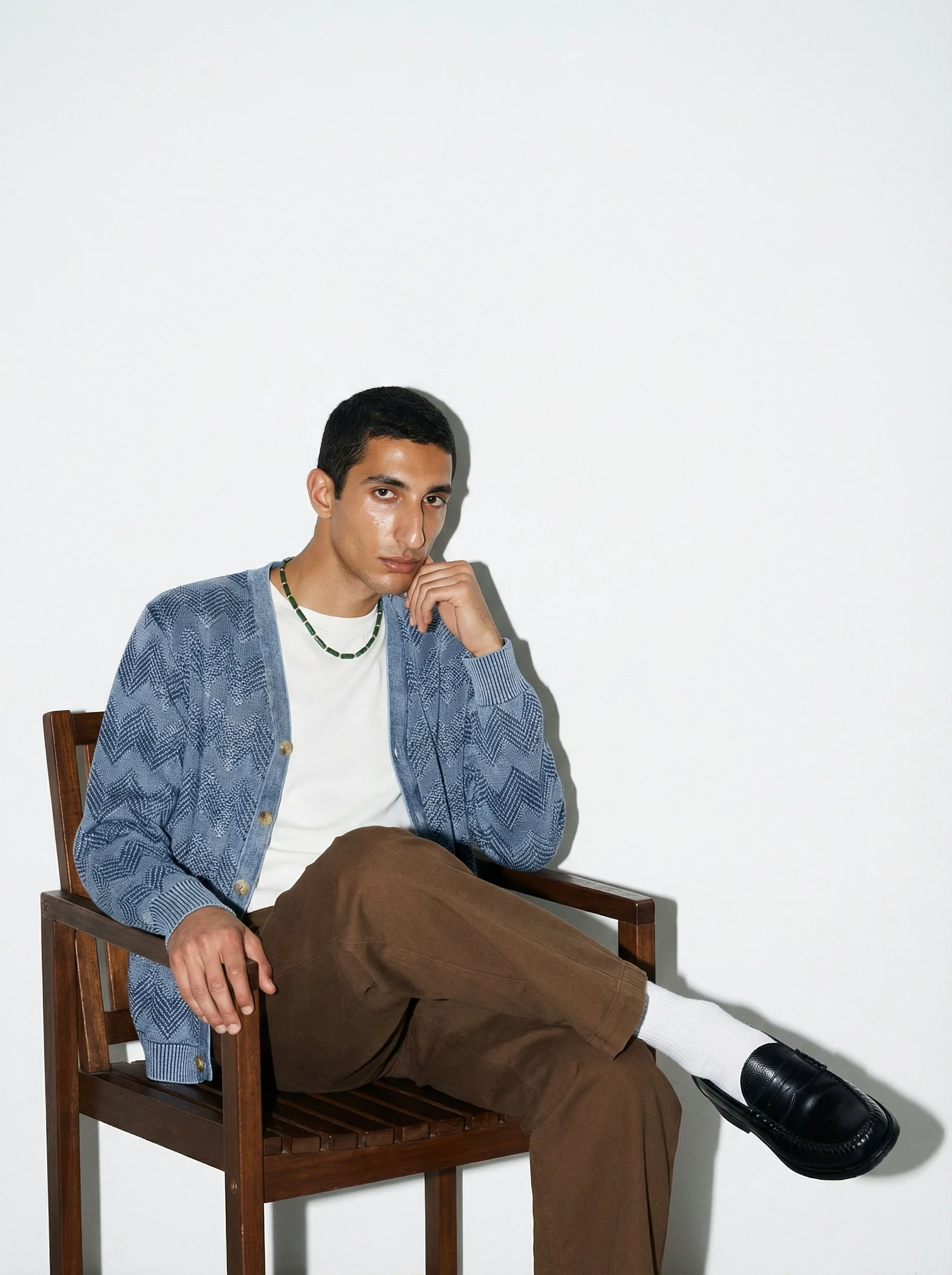 A young man sitting on a wooden chair against a plain white background, wearing a blue patterned cardigan over a white T-shirt, brown pants, and black shoes, with one leg crossed over the other and resting his hand on his chin, looking thoughtfully a