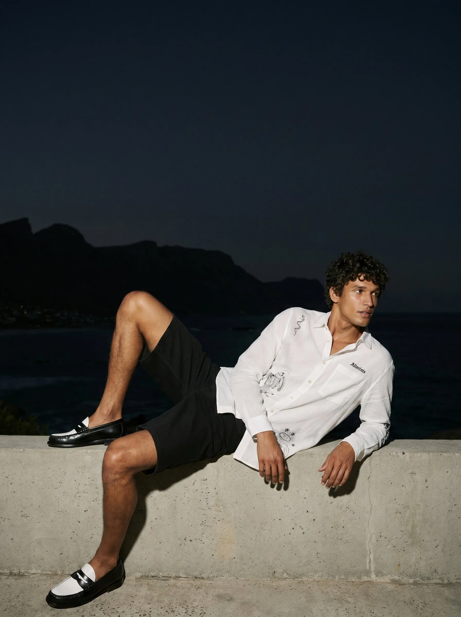 A young man with curly hair relaxing on a concrete ledge at night, dressed in a white shirt with black shorts and black-and-white loafers, with a mountain range in the background under a dark sky.