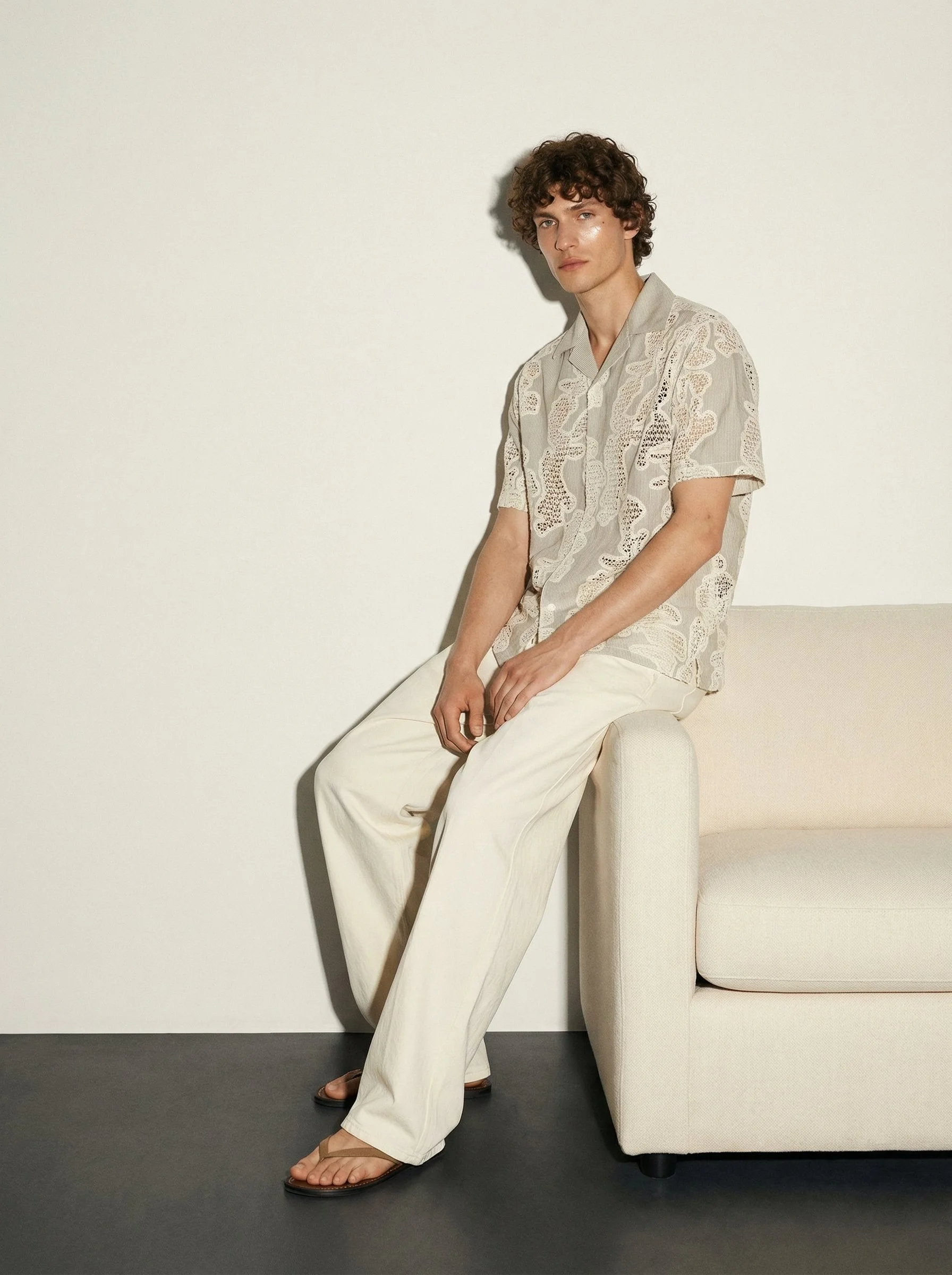 A young man with curly brown hair is sitting on the arm of a beige sofa against a plain white wall. He is wearing a light patterned short-sleeve shirt and cream-colored wide-leg pants, and is wearing brown sandals.