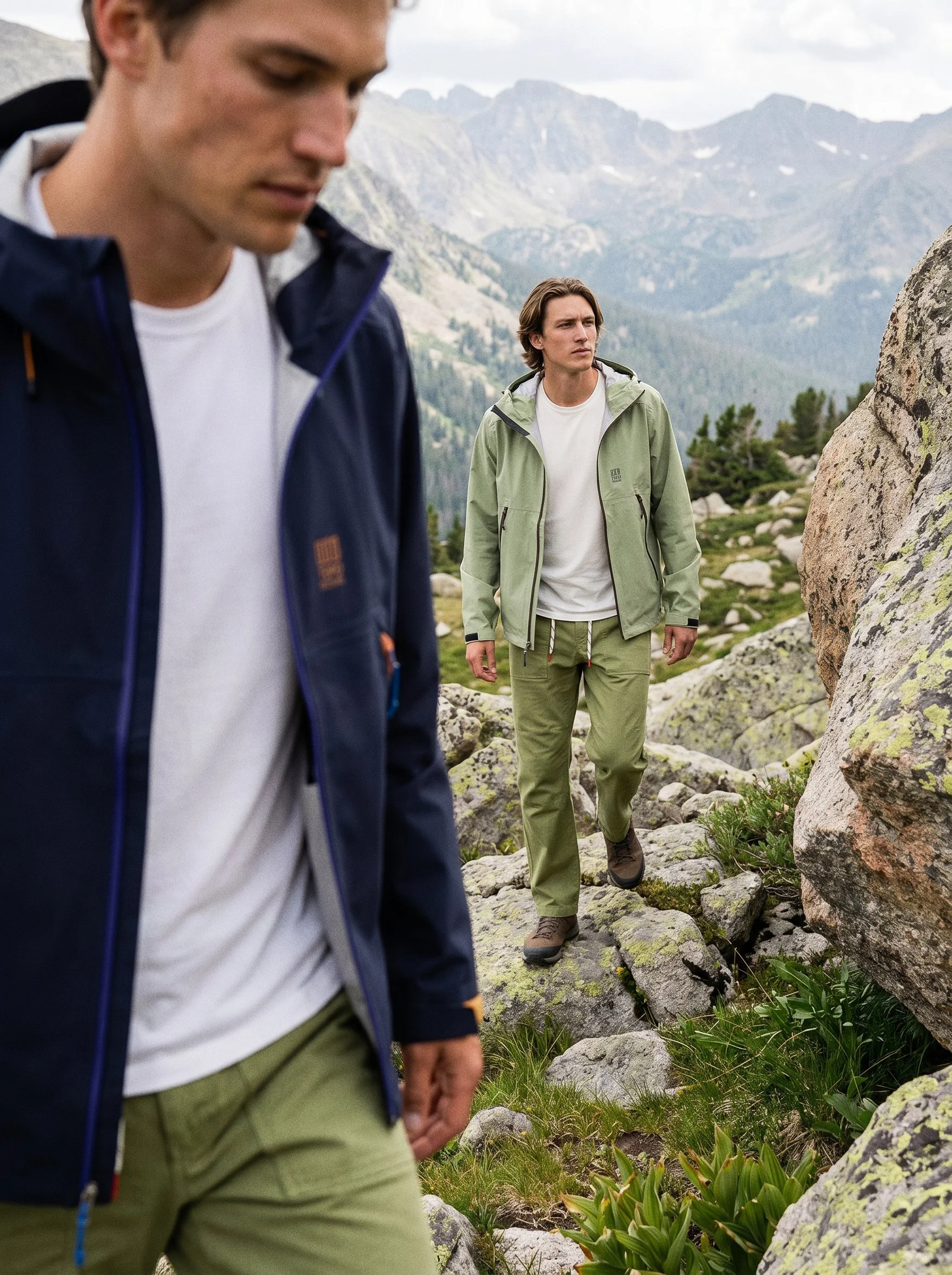 Two young men hiking on rocky trail in mountainous landscape with green vegetation and cloudy sky.