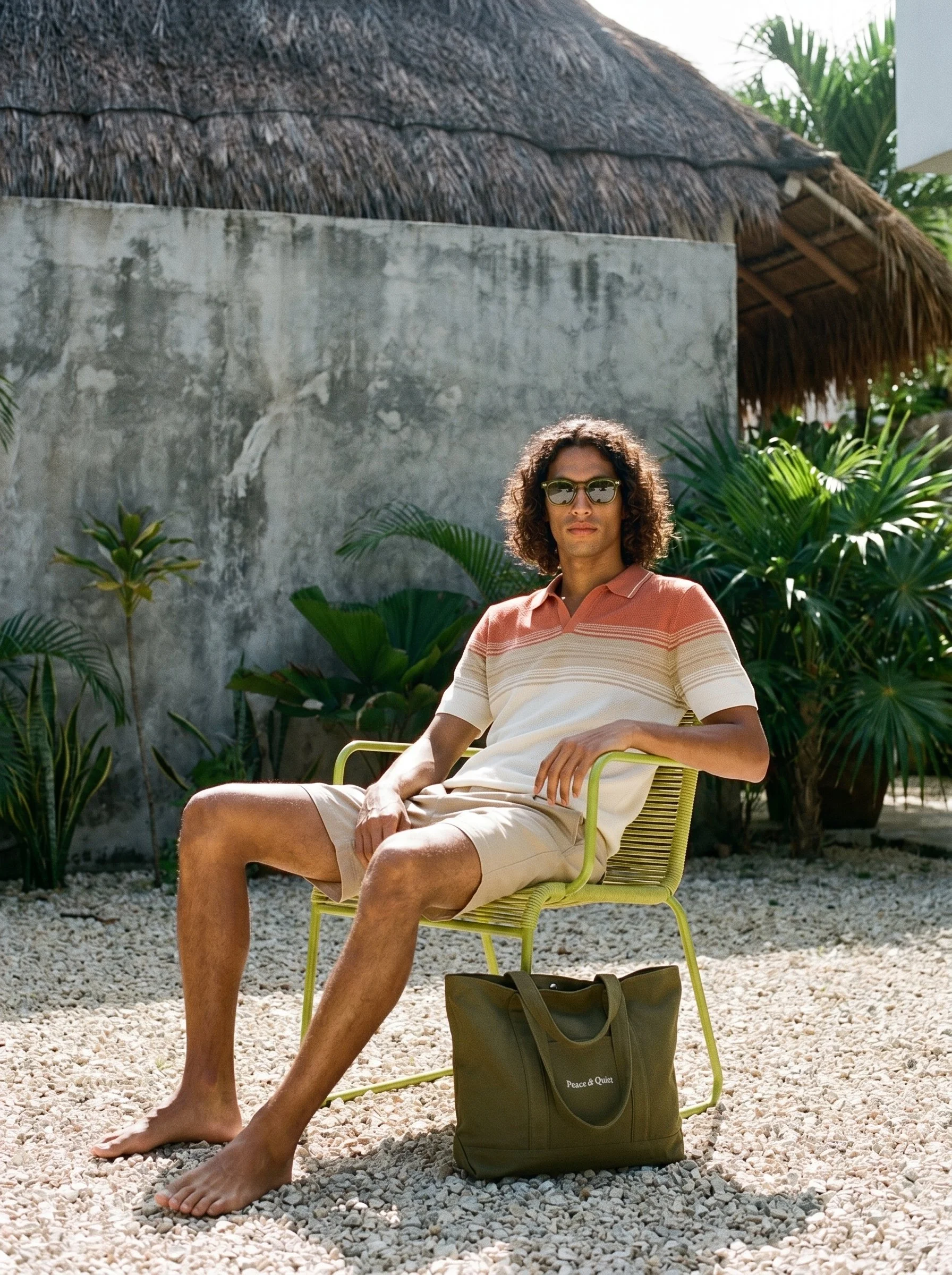 A man with curly hair wearing sunglasses and a striped polo shirt, sitting outdoors on a green chair on a gravel surface, with lush plants, a concrete wall, and a thatched roof structure in the background.