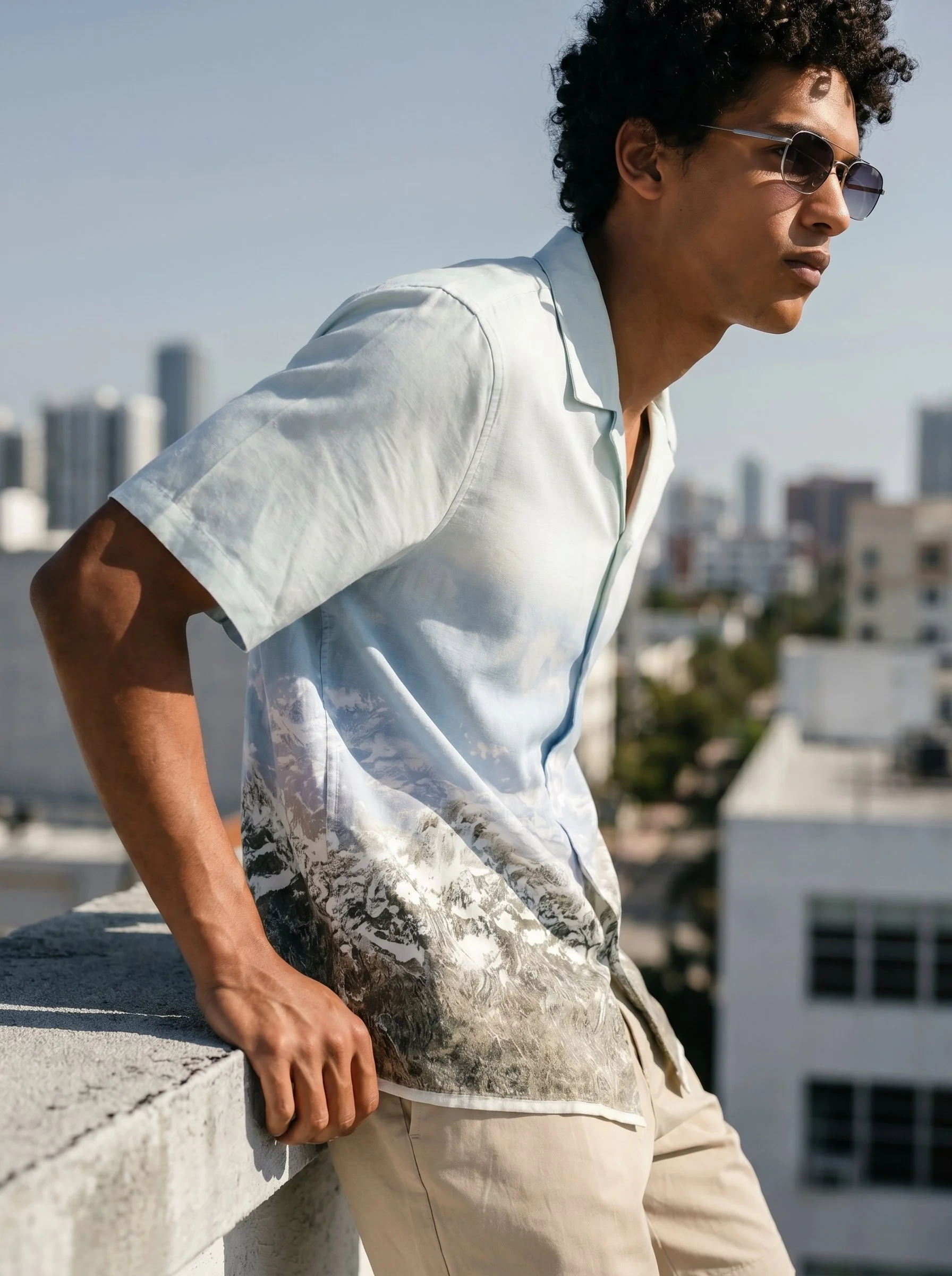 Young man with curly hair wearing sunglasses, a light blue short-sleeve shirt, and beige pants, leaning on a concrete railing on a city rooftop with buildings in the background.