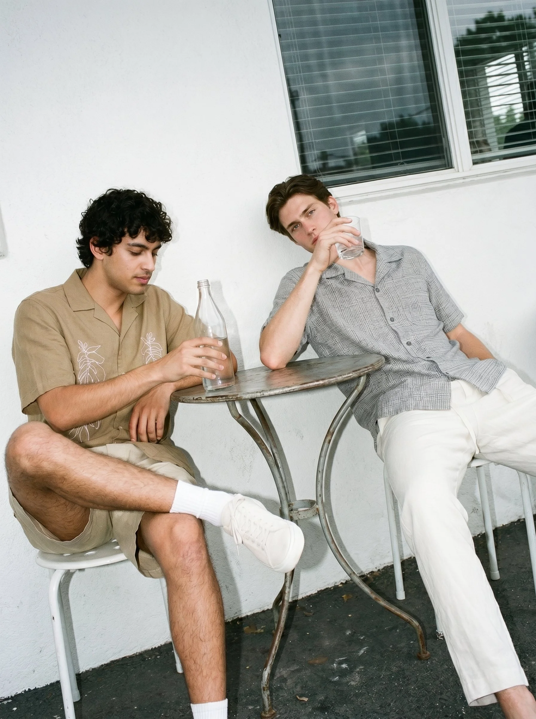 Two young men sitting outside near a white wall and window, one looking down and the other drinking water from a glass.