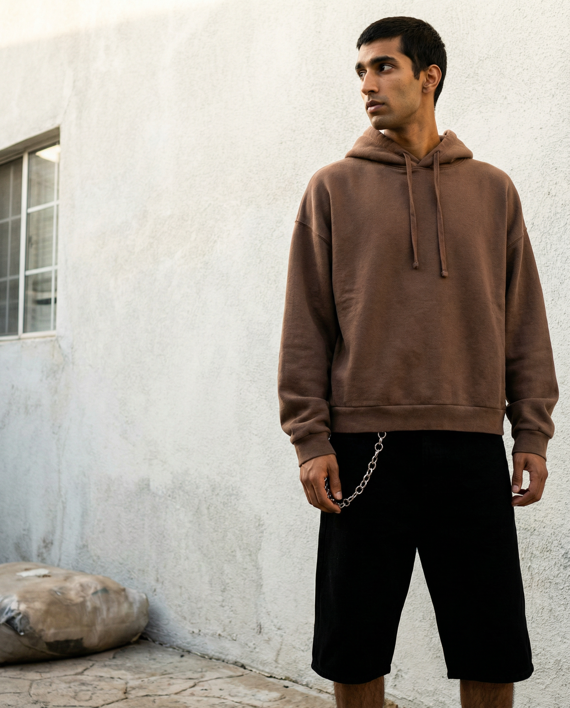 Young man standing outdoors against a white wall, wearing a brown hoodie and black shorts with a silver chain attached to his waistband.  AI fashion editorial.