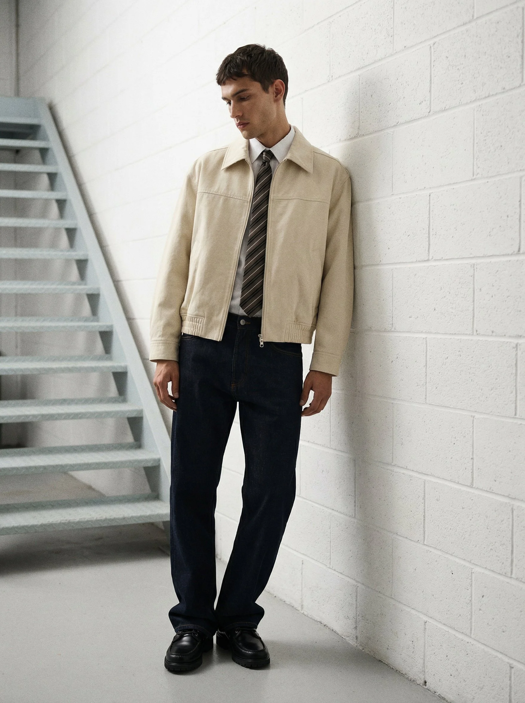 Young man in beige jacket, white shirt, striped tie, dark pants, and black shoes standing against a white brick wall next to a staircase.  AI fashion editorial.