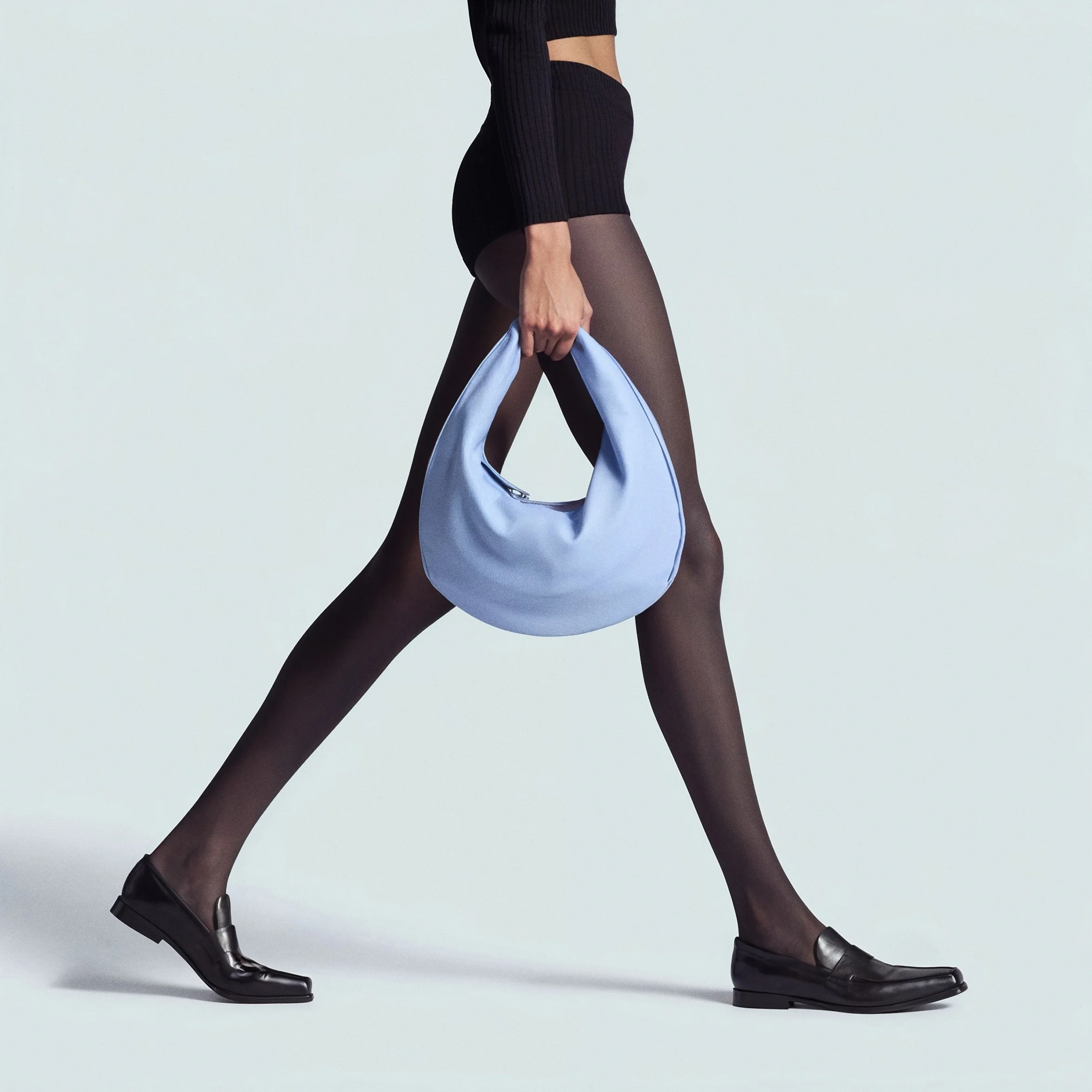 Lower half of a person walking, wearing black shoes, black tights, a black skirt, and a black long-sleeve top, carrying a light blue handbag.