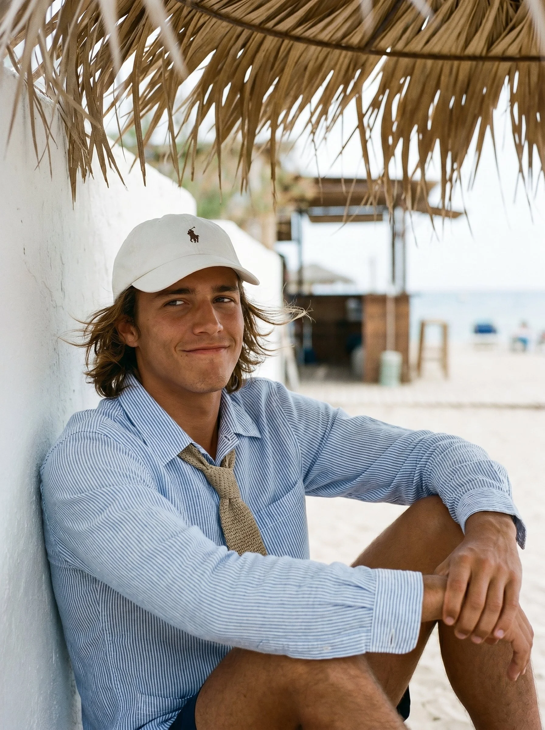 Young man sitting on a beach, leaning against a white wall under a thatched roof, smiling, with a beach scene in the background.