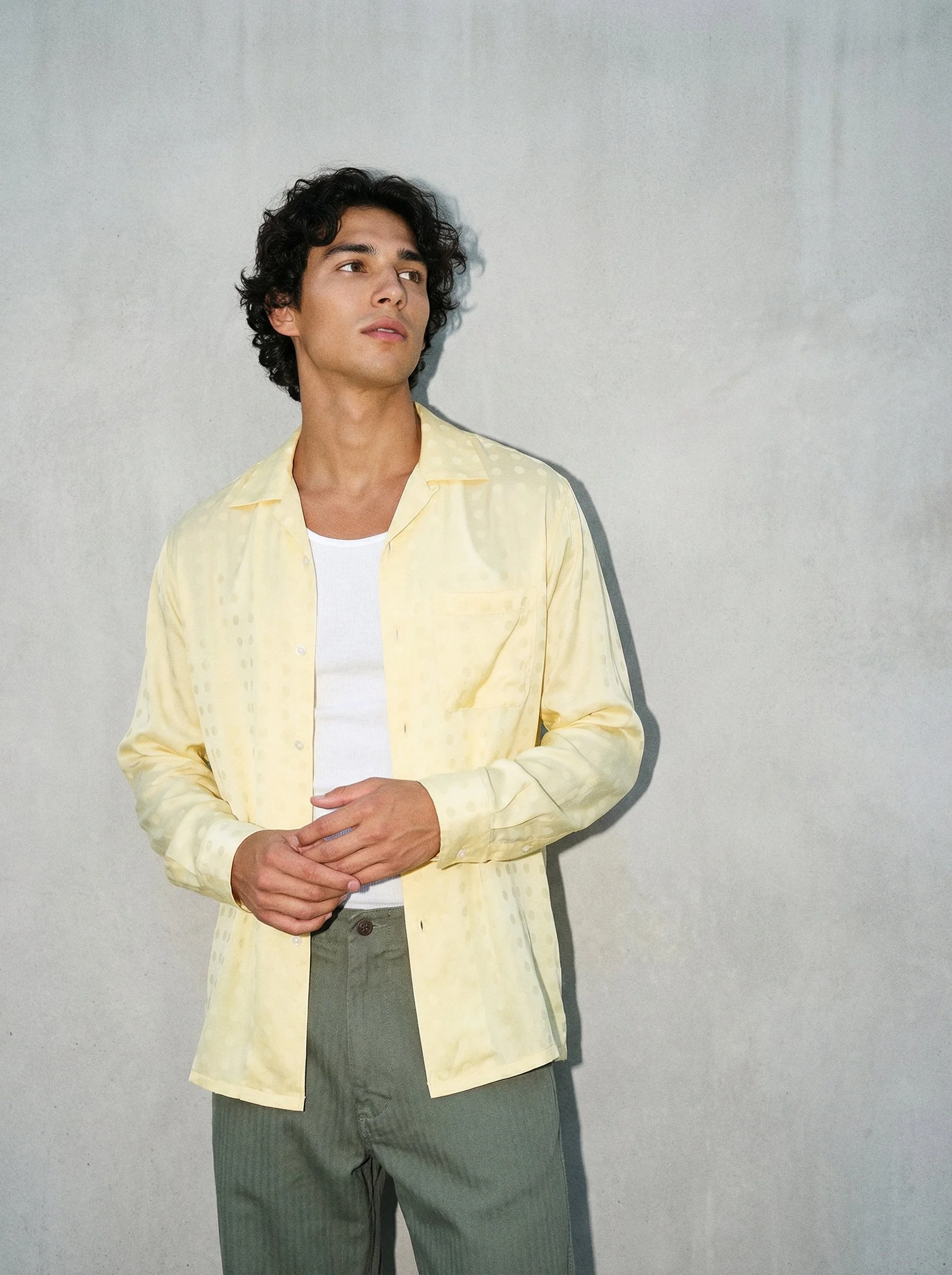 A young man with curly dark hair wearing a light yellow button-up shirt over a white T-shirt, standing against a plain gray wall, with his hands gently clasped at his waist, looking to the side.  AI fashion editorial.
