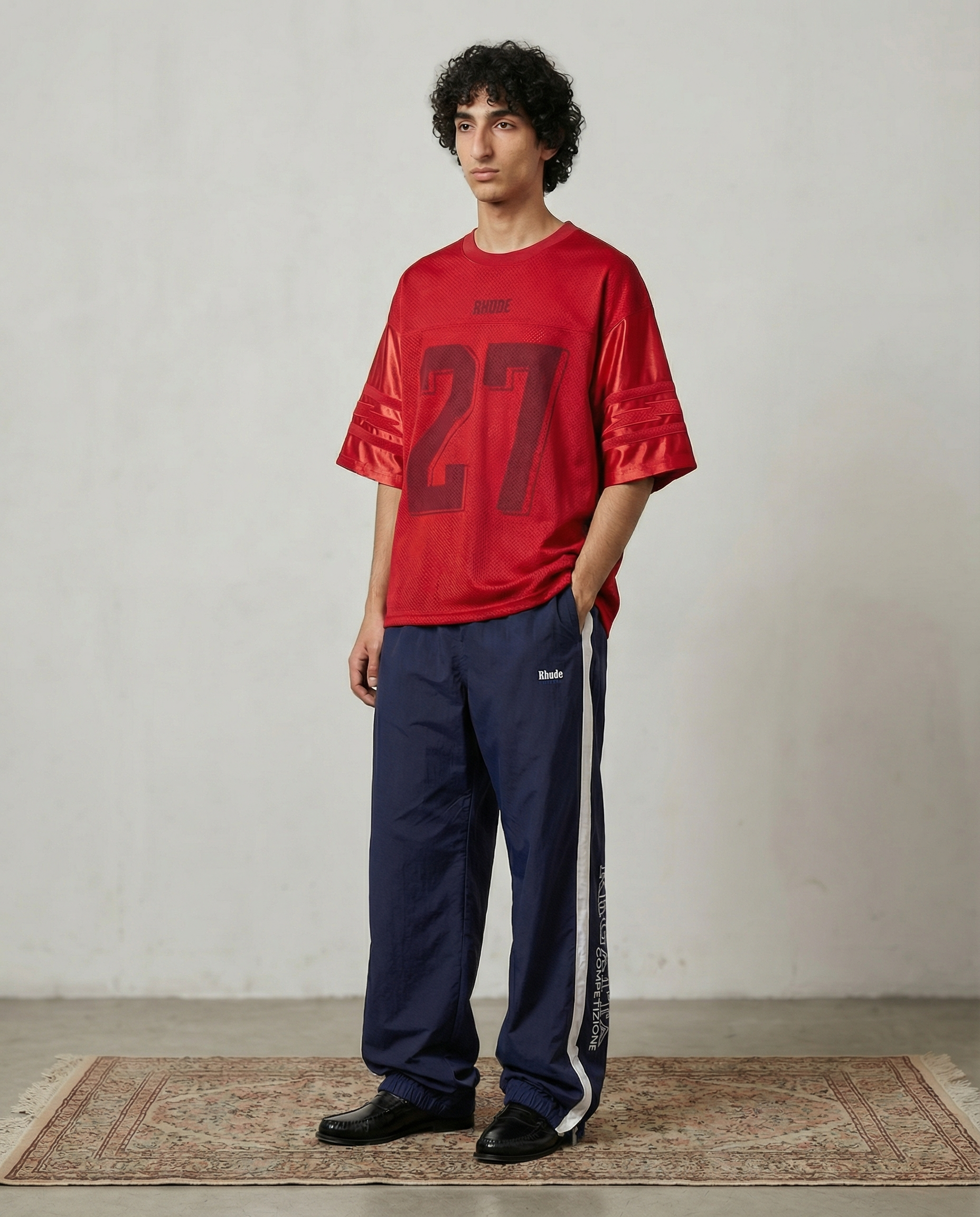 A young man with curly dark hair stands on a patterned area rug against a plain light gray wall. He is wearing a red sports jersey with the number 22 and the word "RHODE" on it, navy blue athletic pants with a white stripe down the side and a logo, a