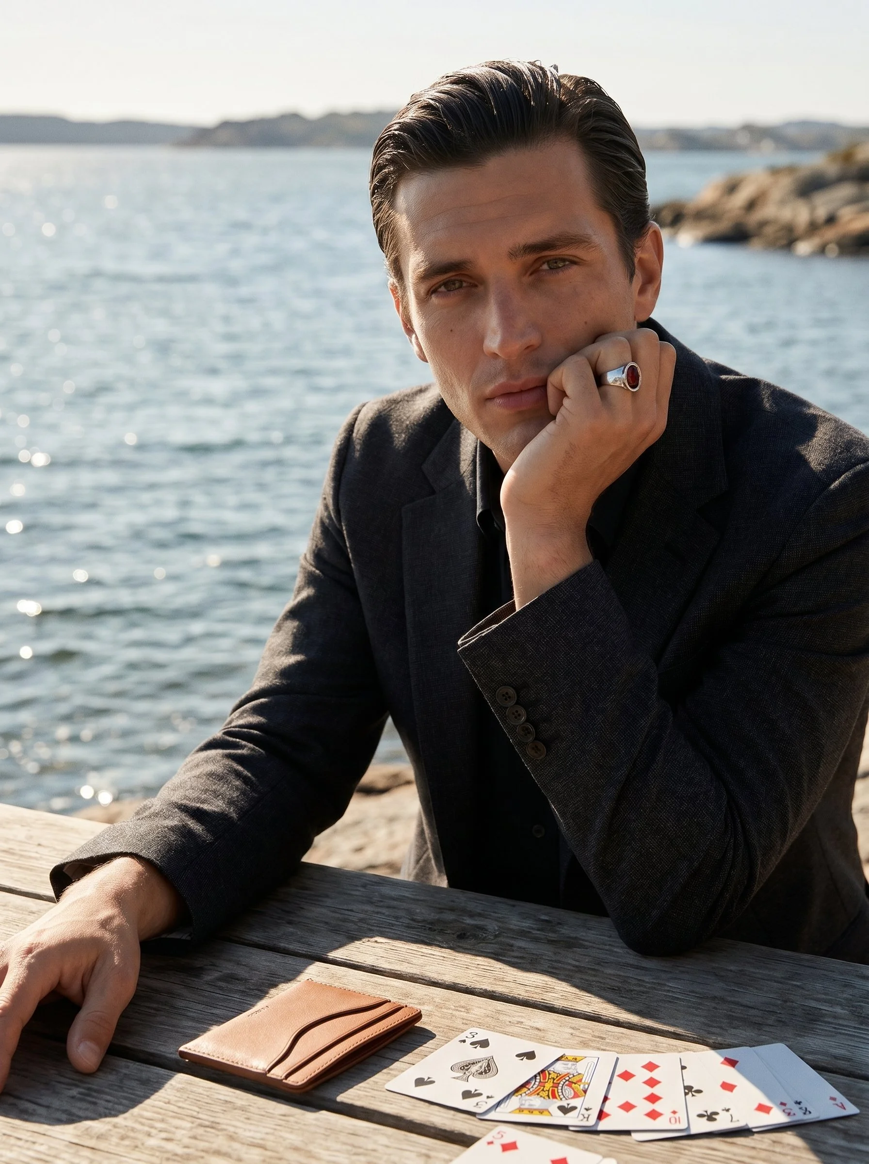 A young man with dark hair and wearing a dark blazer rests his chin on his hand, looking at the camera, with a body of water and distant land in the background. On a wooden table in front of him are playing cards, a pink wallet, and a person's finger