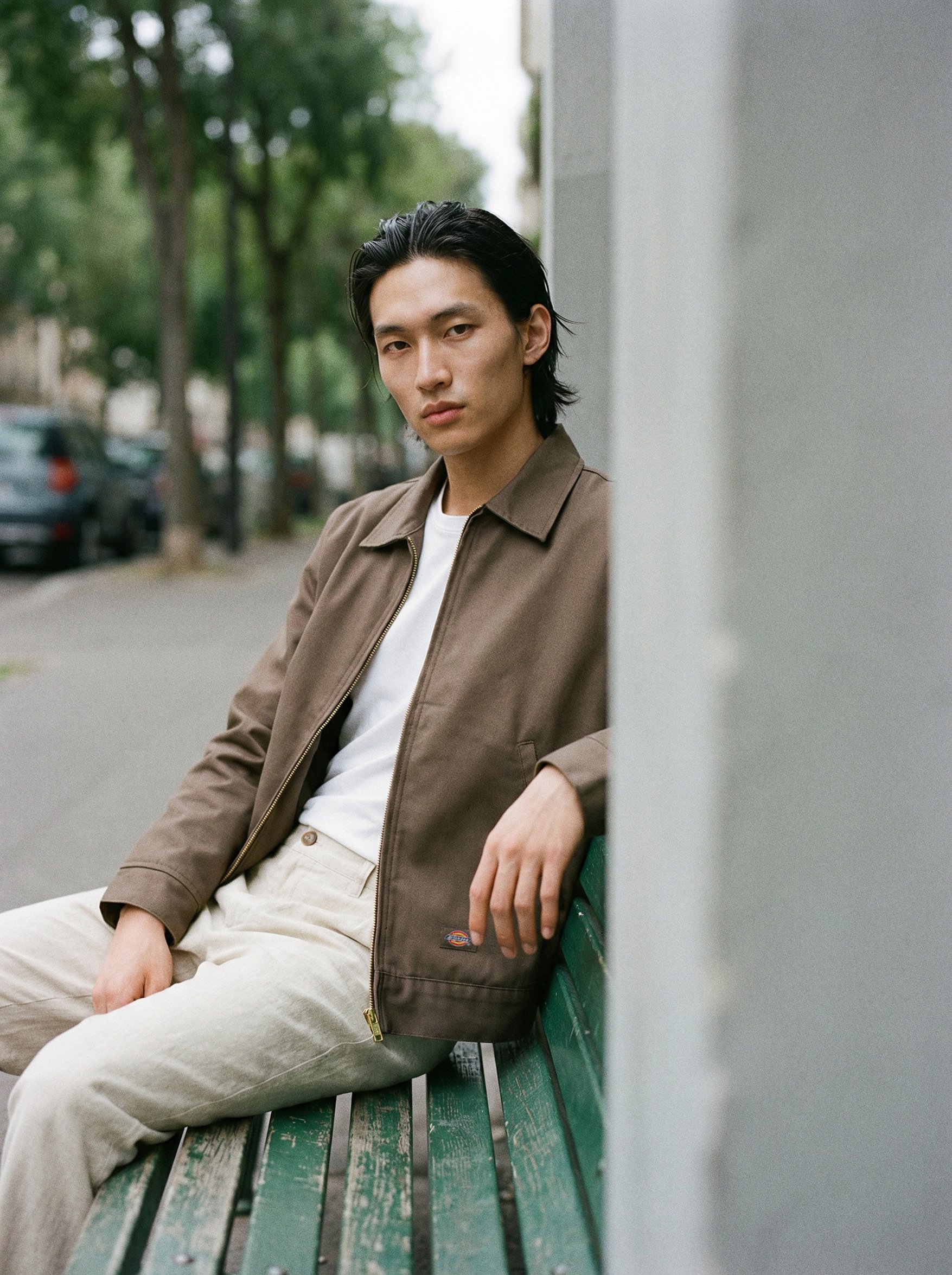 A young man with dark, slicked-back hair sitting on a green park bench next to a wall, outdoors with trees in the background, wearing a brown jacket over a white shirt and beige pants.  AI fashion editorial.