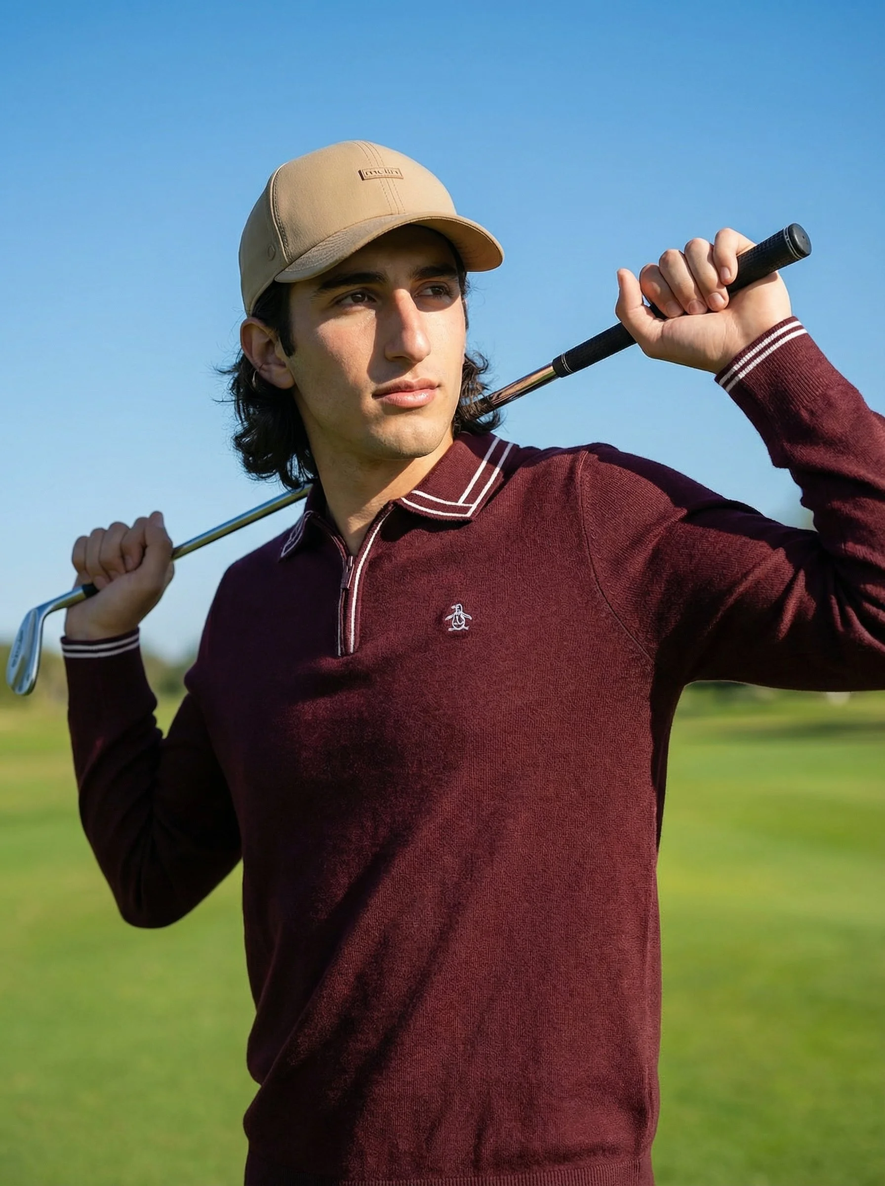 A young man wearing a beige cap and a maroon zip-up sweater holding a golf club over his shoulder on a golf course.