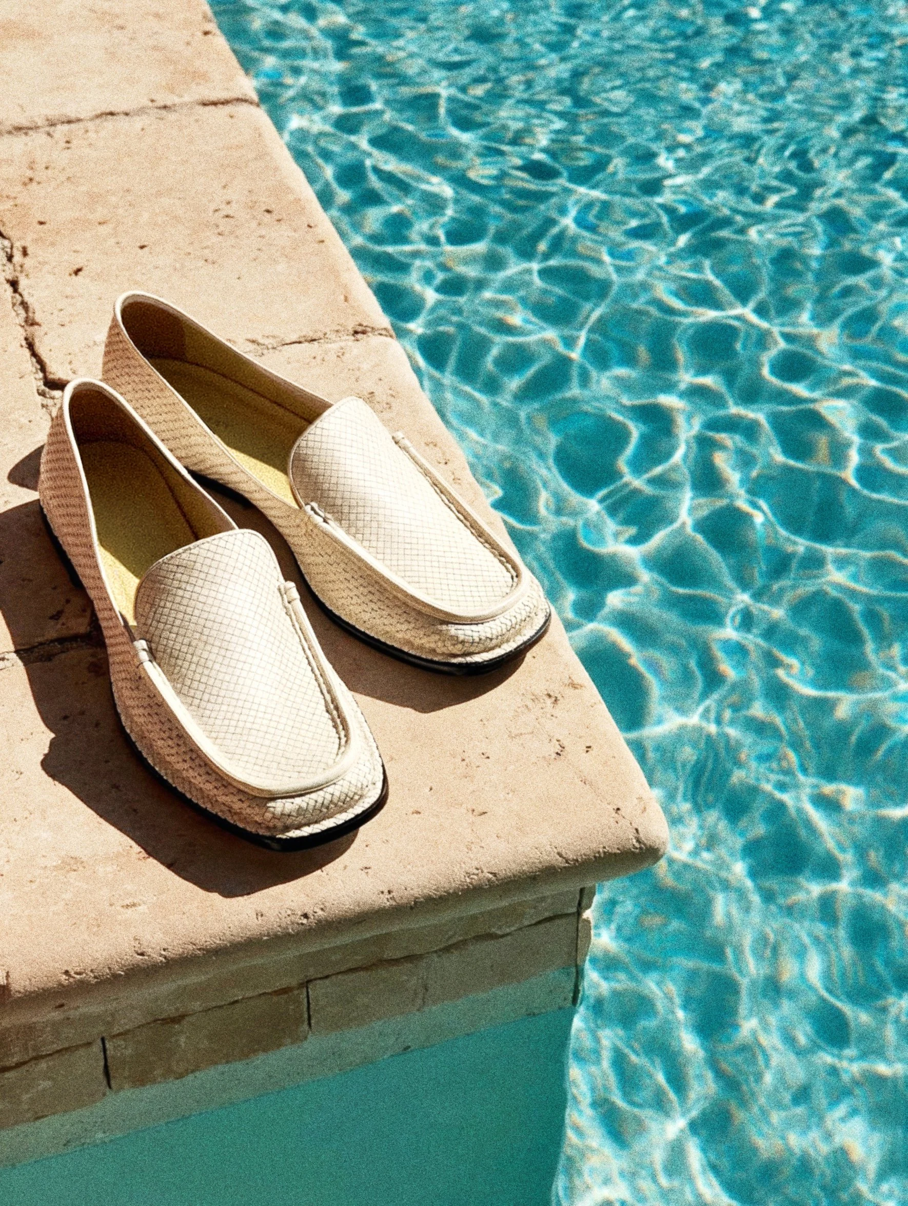 Pair of white slip-on shoes resting on the edge of a swimming pool with clear blue water.