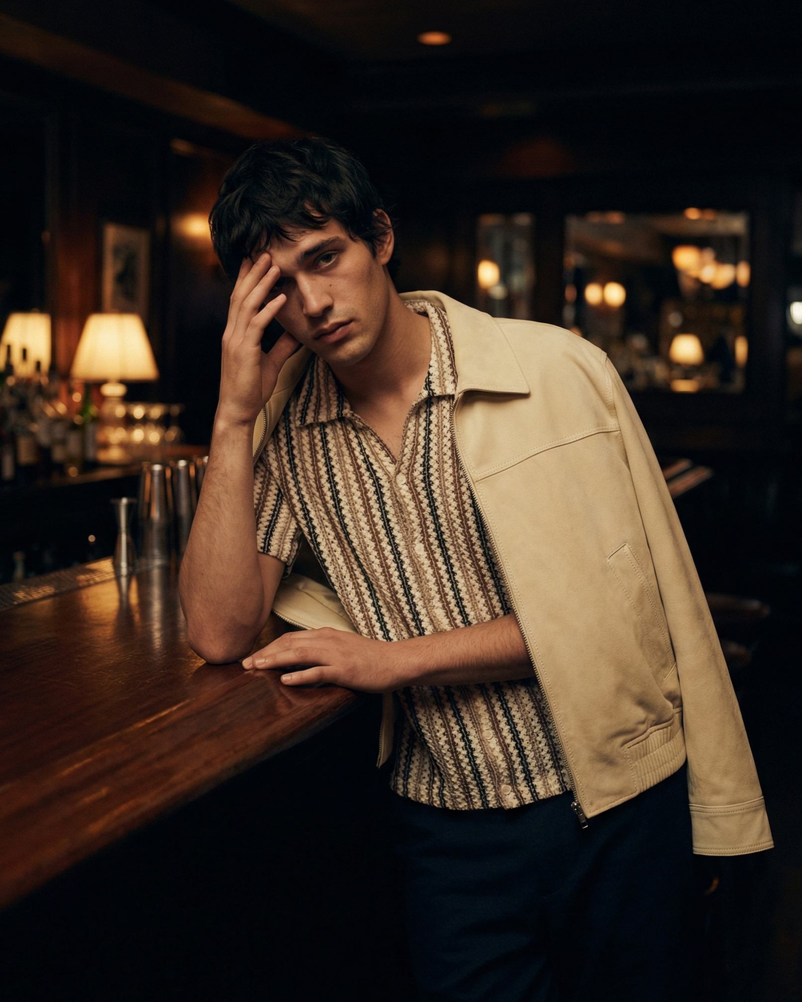 Young man with dark hair resting his head on his hand at a bar or cafe, with warm lighting and blurred background.