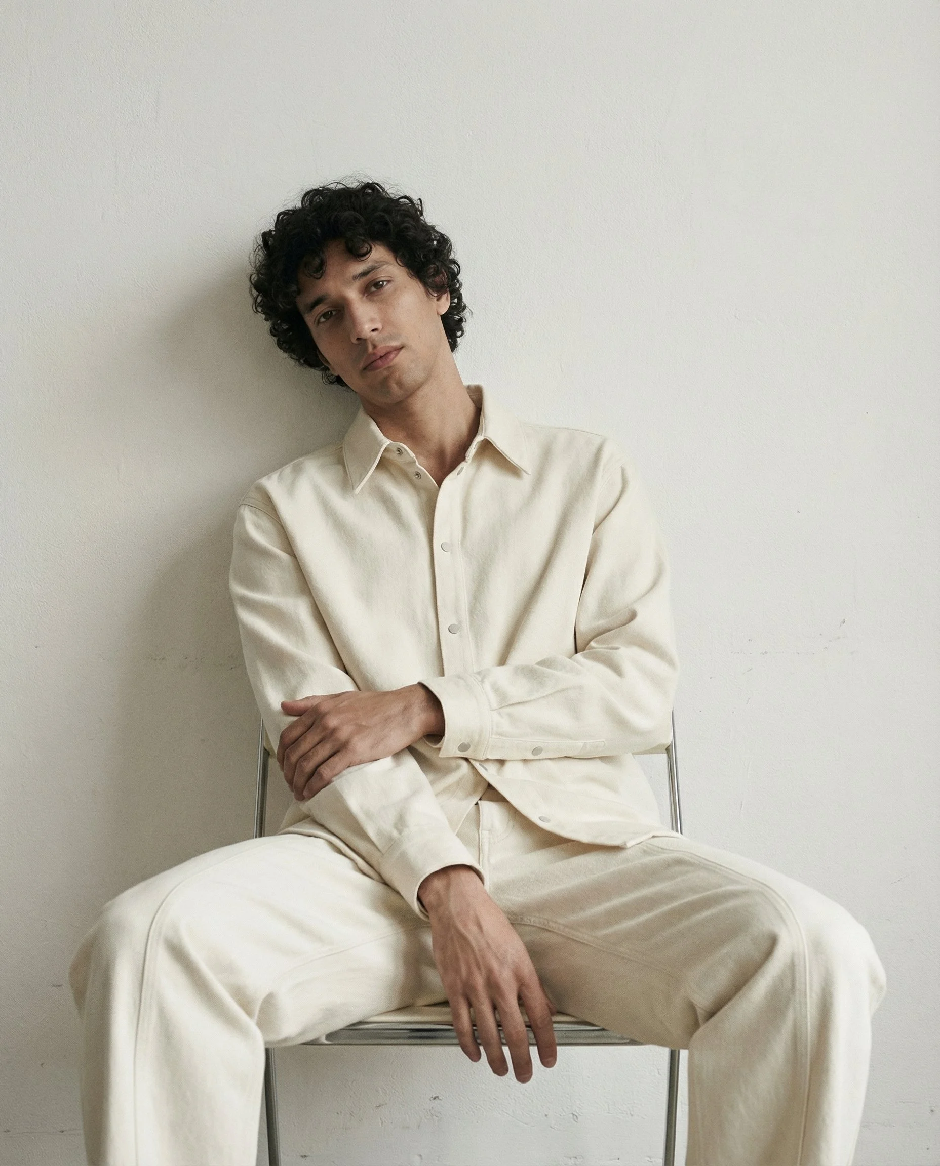 A young man with curly dark hair, wearing a cream-colored shirt and pants, sitting on a chair against a plain white wall. His head is slightly tilted, with a calm expression, arms crossed, and left hand resting on his lap.