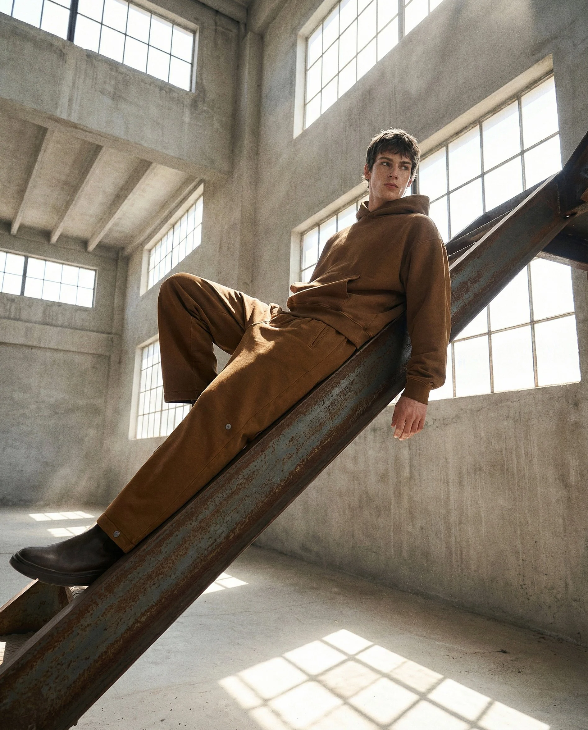 A young man sitting on a rusty metal slide inside an industrial-style building with large windows and concrete walls, sunlight casting grid-like shadows on the floor.