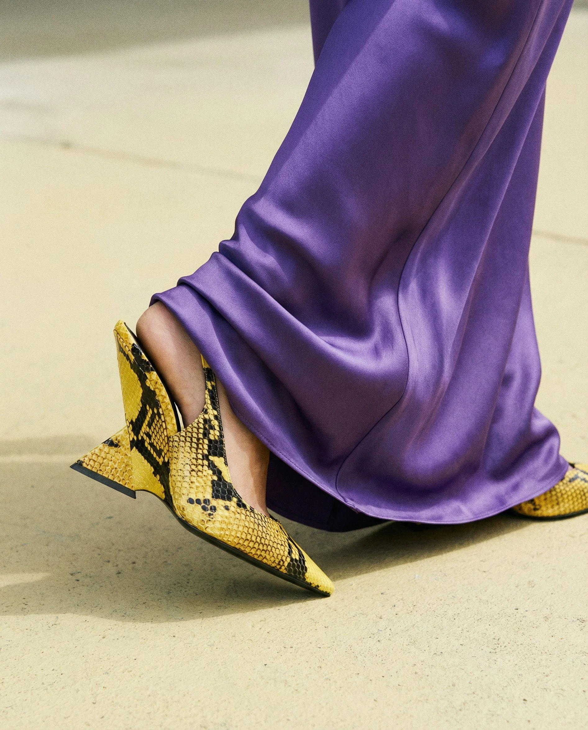 Close-up of a person's leg wearing purple satin pants and yellow snakeskin shoes, sitting on a beige floor.