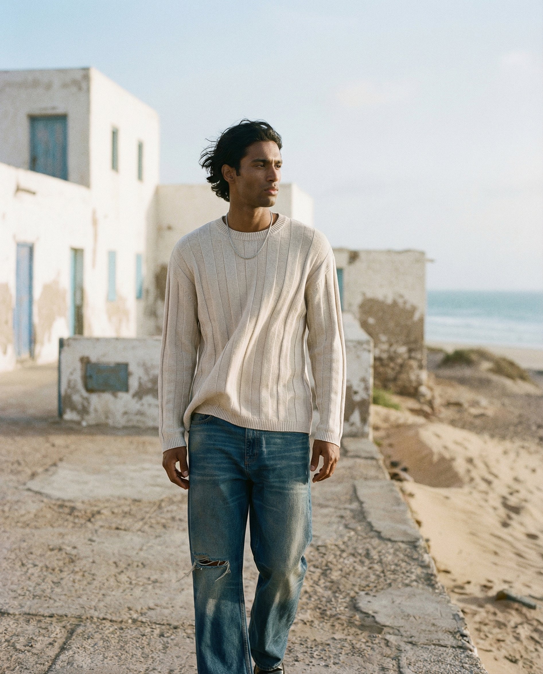 A young man with black hair, wearing a beige sweater and distressed jeans, stands outdoors on a concrete pathway in front of white buildings by the sea during daylight.