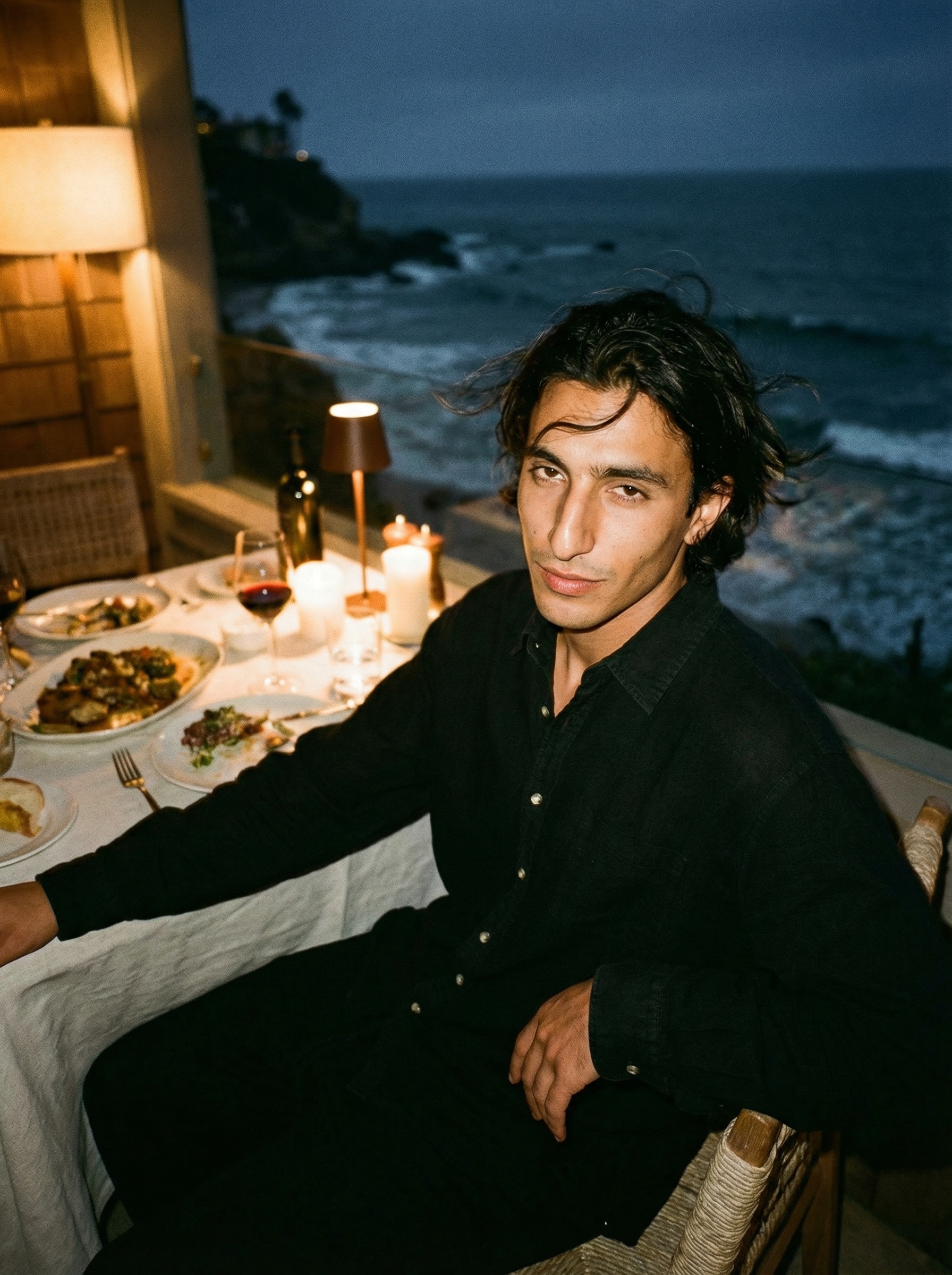 A young man with dark, slightly wavy hair and a black shirt is sitting at a dinner table by the coast during evening or night. The table has various dishes and candles, and the ocean is visible in the background with dark waves and a cloudy sky.