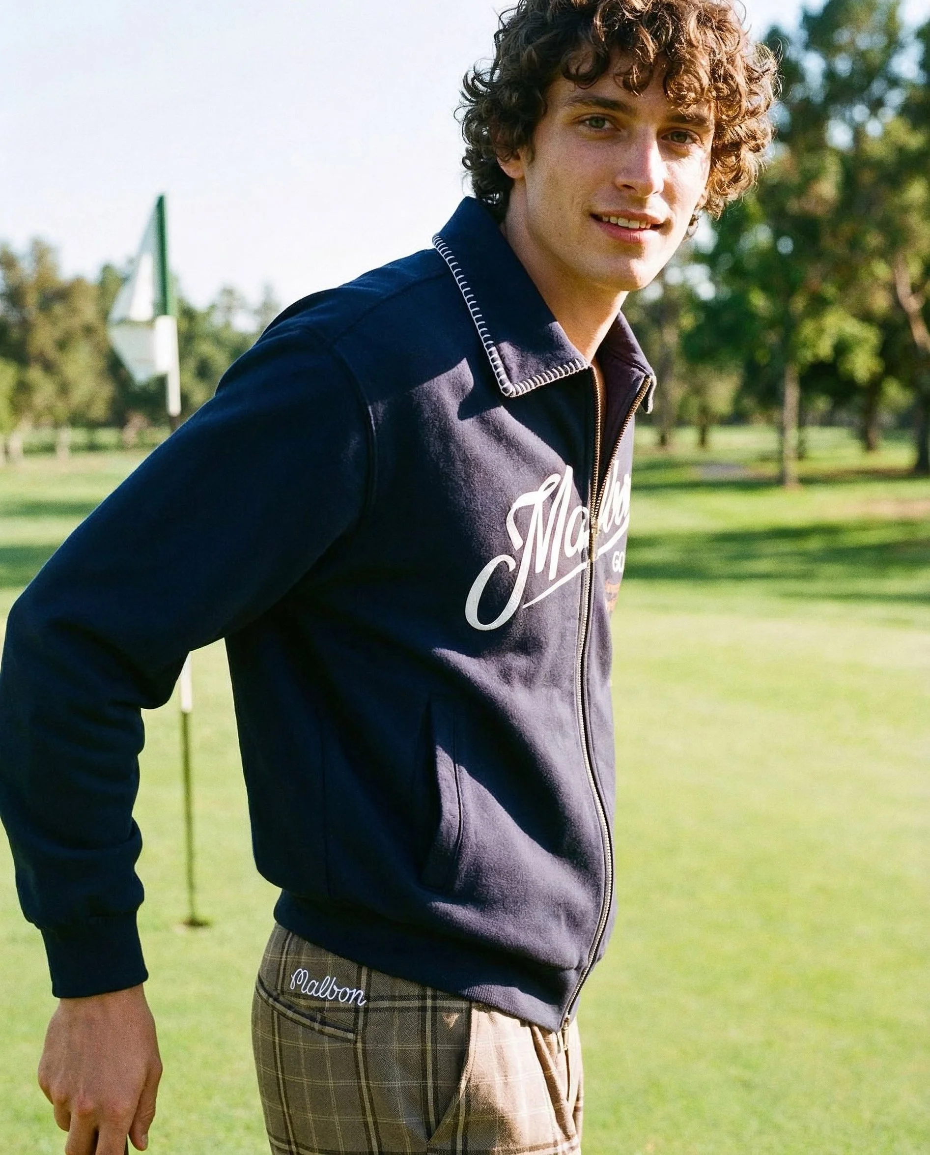 A young man standing outdoors on a grassy field, with trees and a flag in the background. He has curly hair, is wearing a dark blue zip-up hoodie with white lettering, and plaid shorts.