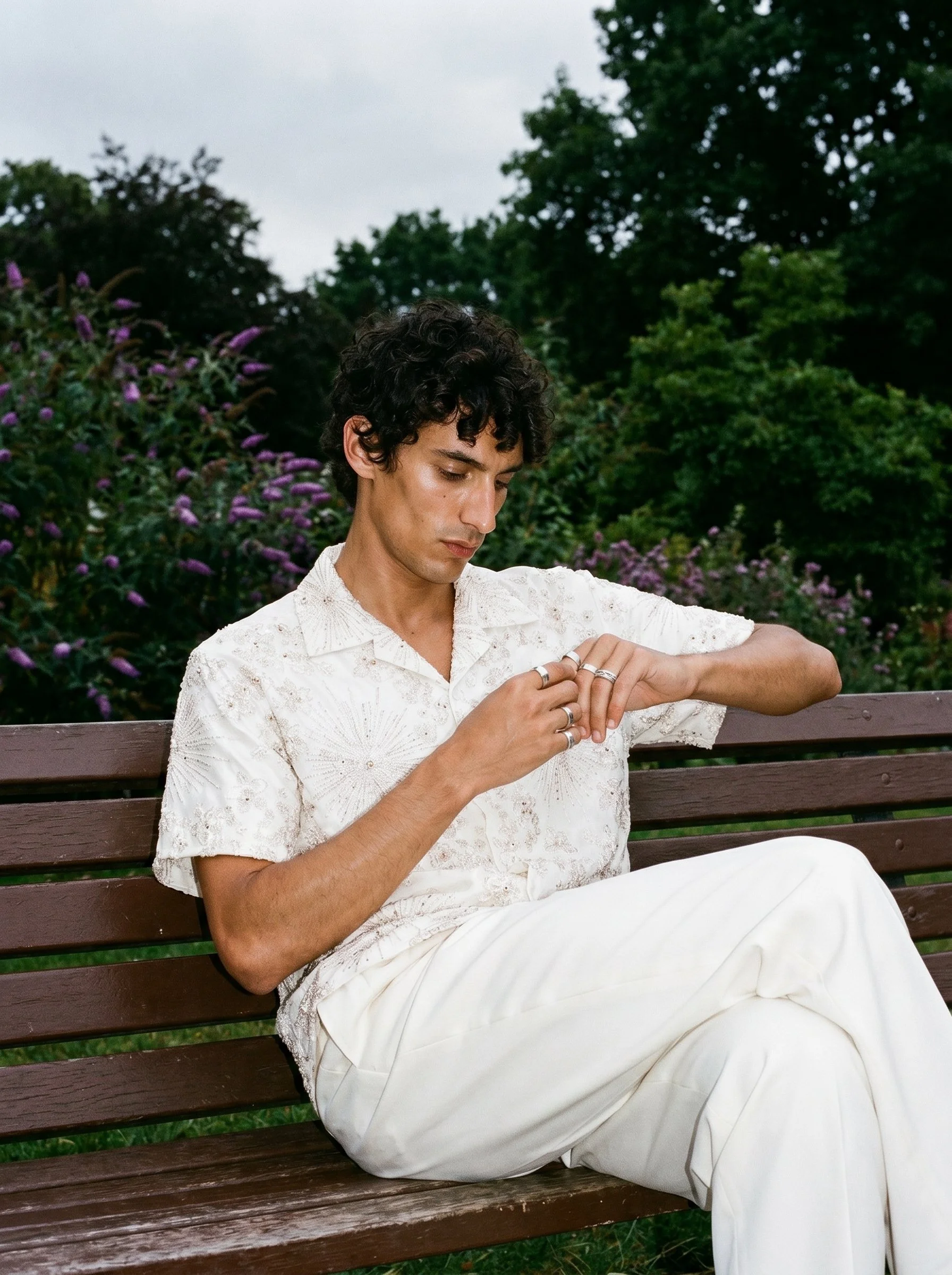 A young man with curly dark hair sitting on a park bench during daytime, wearing a white embroidered shirt and white pants, looking down and adjusting rings on his fingers, with green trees and purple flowering bushes in the background.