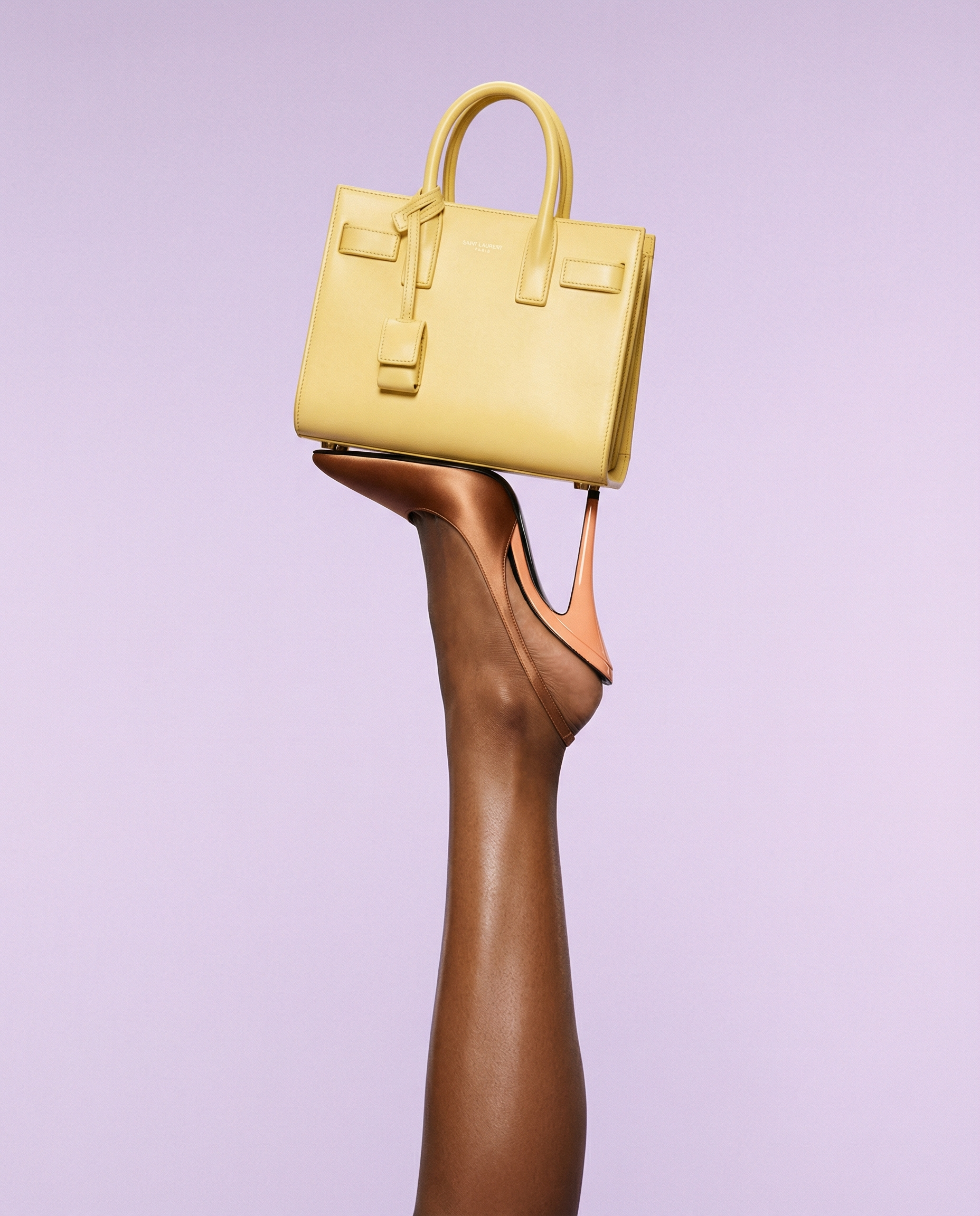 A yellow handbag with a structured shape and top handles, suspended in the air above a high-heeled shoe against a light purple background.