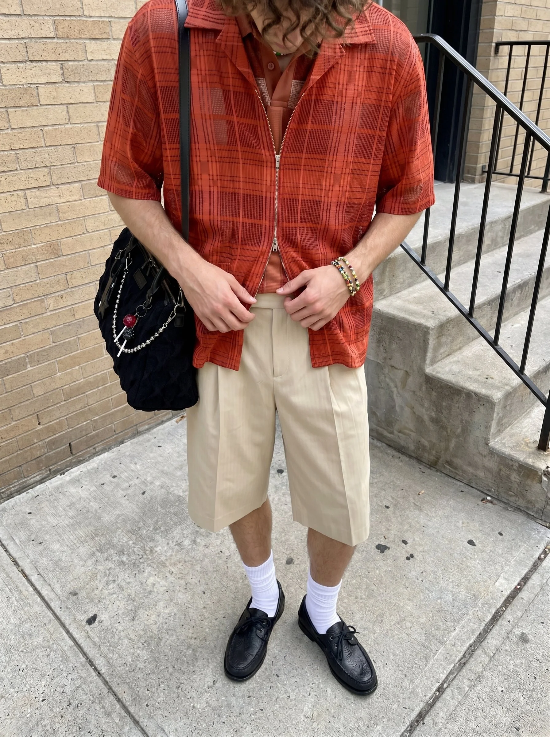 Person standing outdoors in front of a brick wall and concrete stairs, wearing beige shorts, white socks, black shoes, a red plaid jacket, beige shirt, and multiple beaded bracelets, carrying a black bag with a chain strap.