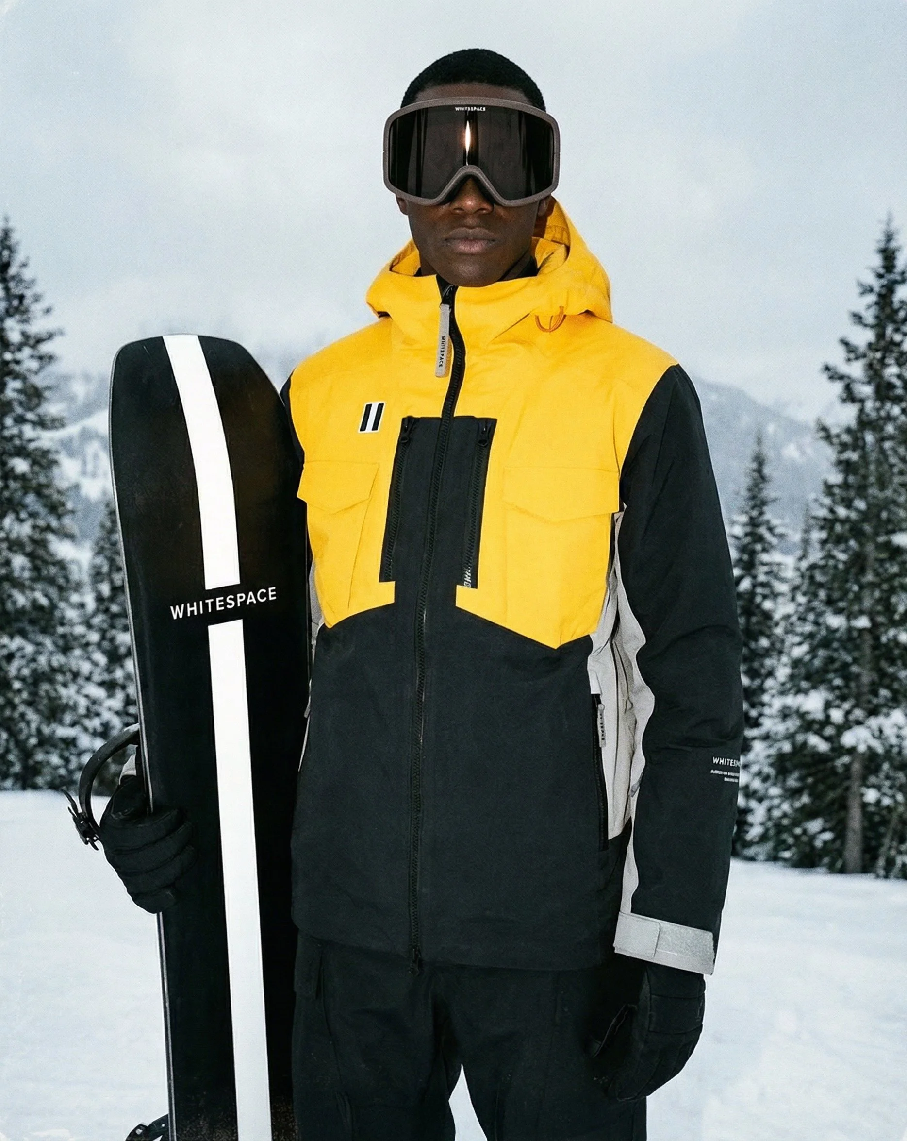 A man in a yellow and black ski jacket wearing ski goggles, holding a snowboard, standing on a snowy landscape with trees in the background.