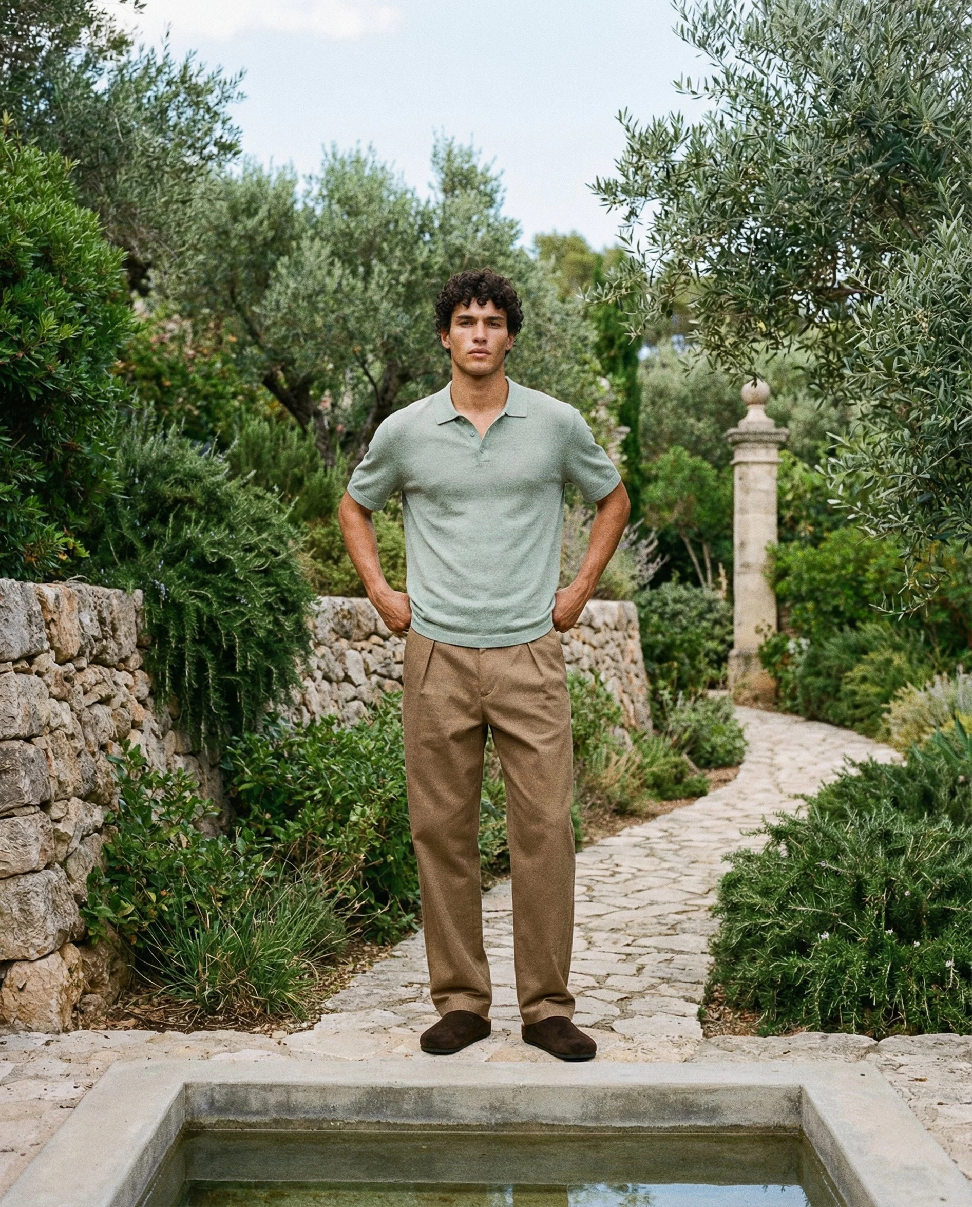 A young man with curly dark hair, wearing a gray polo shirt, tan pants, and dark shoes, stands with hands on hips behind a water feature in a lush garden with stone walls, various plants, and trees. AI fashion editorial image.