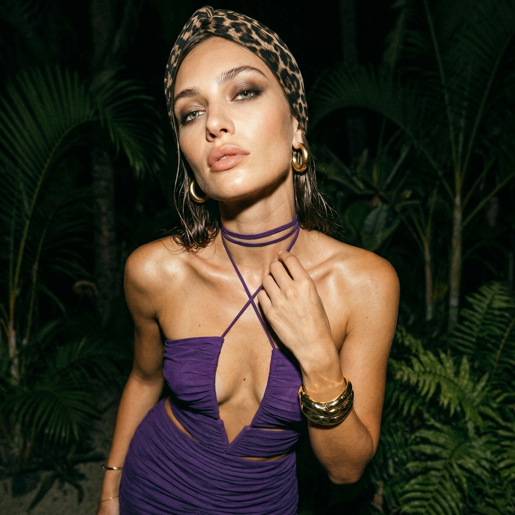 A woman with wavy brown hair wearing a leopard-print headscarf, gold hoop earrings, multiple gold bangles, and a purple strapless dress, standing in front of lush green plants.