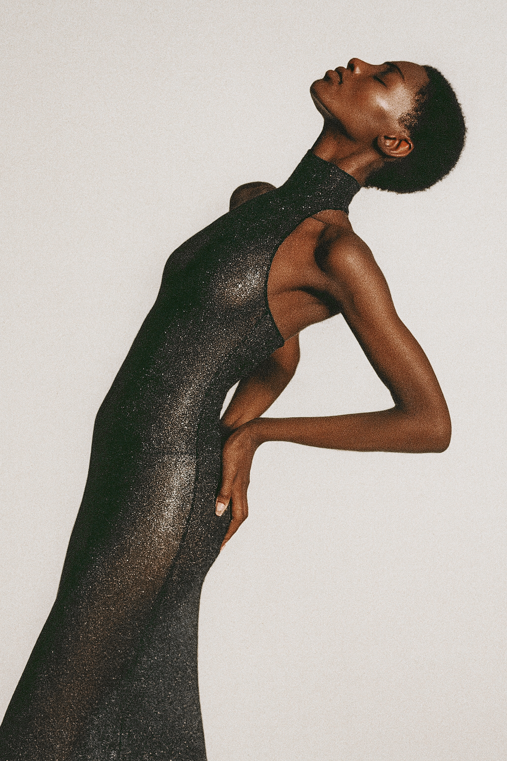A woman with short natural hair poses with her head tilted back and eyes closed, wearing a sparkly black sleeveless dress against a plain light background.  AI fashion editorial image.