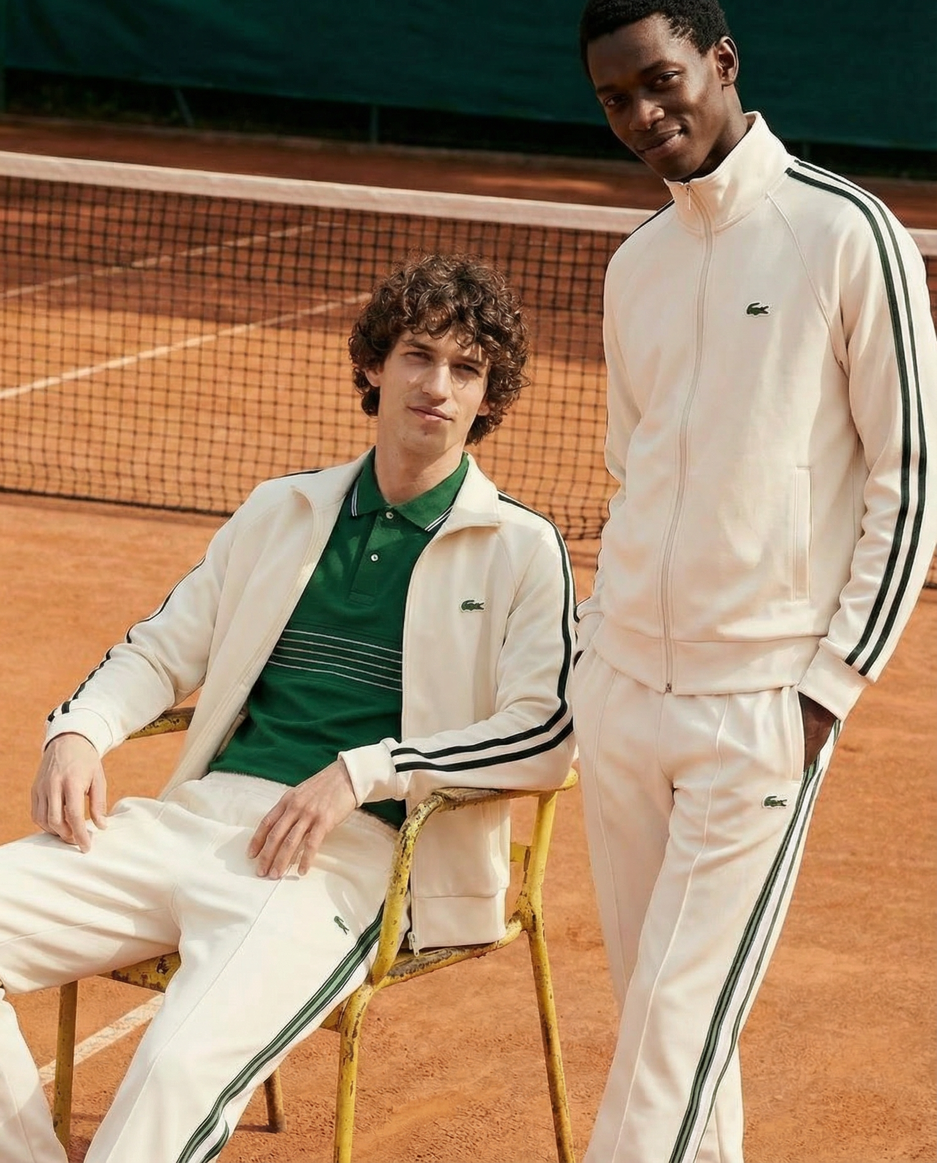 Two young men on a tennis court, one sitting in a yellow chair and the other standing beside him. Both are wearing matching white tracksuits with dark green accents and Lacoste logos. The court surface is reddish, and a net is visible in the backgrou