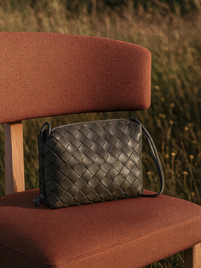 A black woven leather handbag resting on a reddish-brown fabric chair with a wooden frame, outdoors on a grassy field.