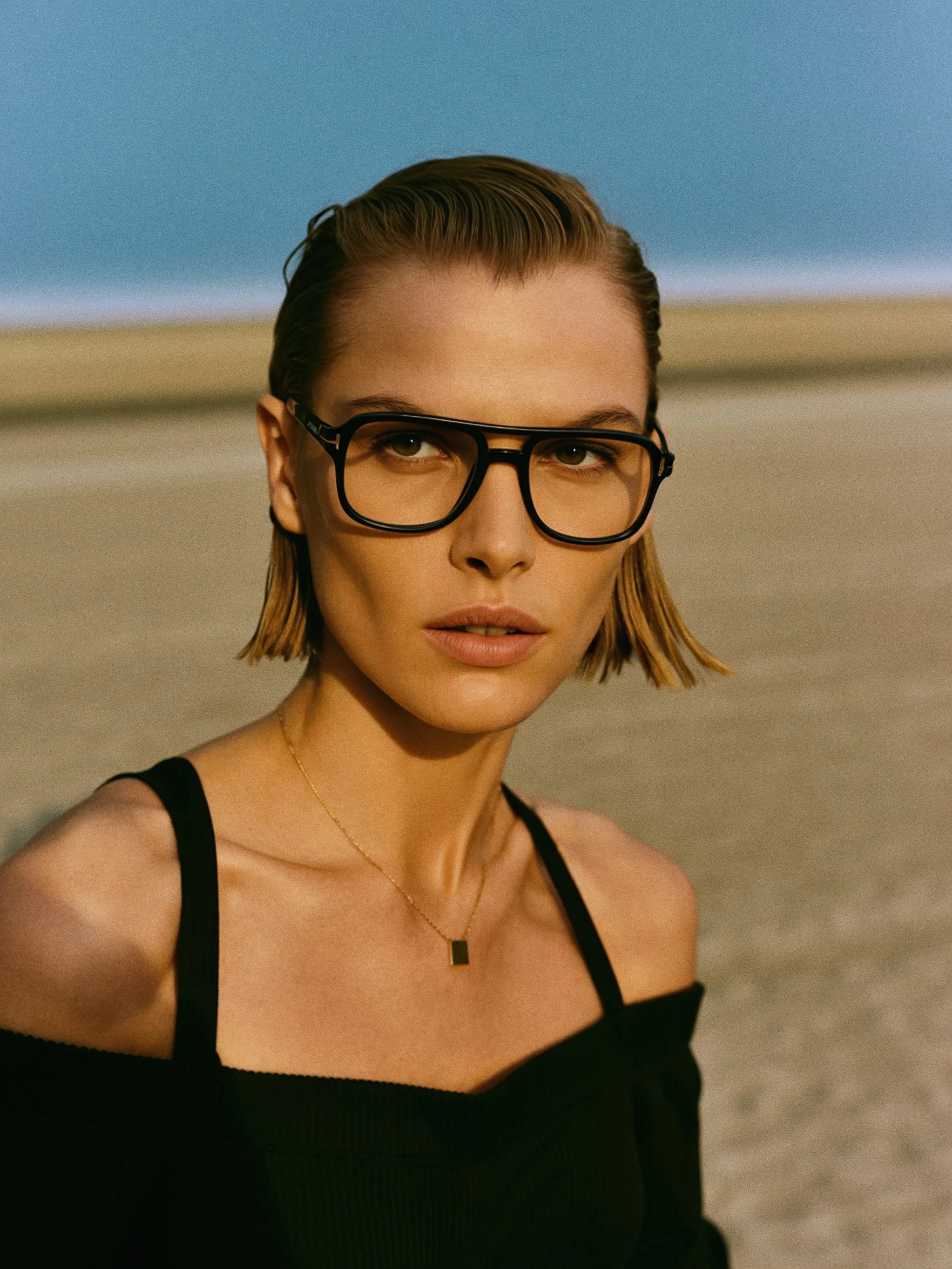 A young woman with short, wet hair wearing black glasses and a black sleeveless top standing outdoors on a beach with sand and a blue sky in the background.