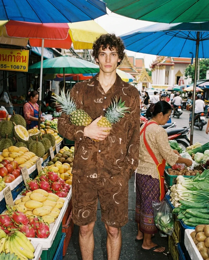 A young man with curly hair wearing a brown patterned outfit, standing in an outdoor marketplace holding two pineapples, surrounded by colorful fruits and market vendors under umbrellas.