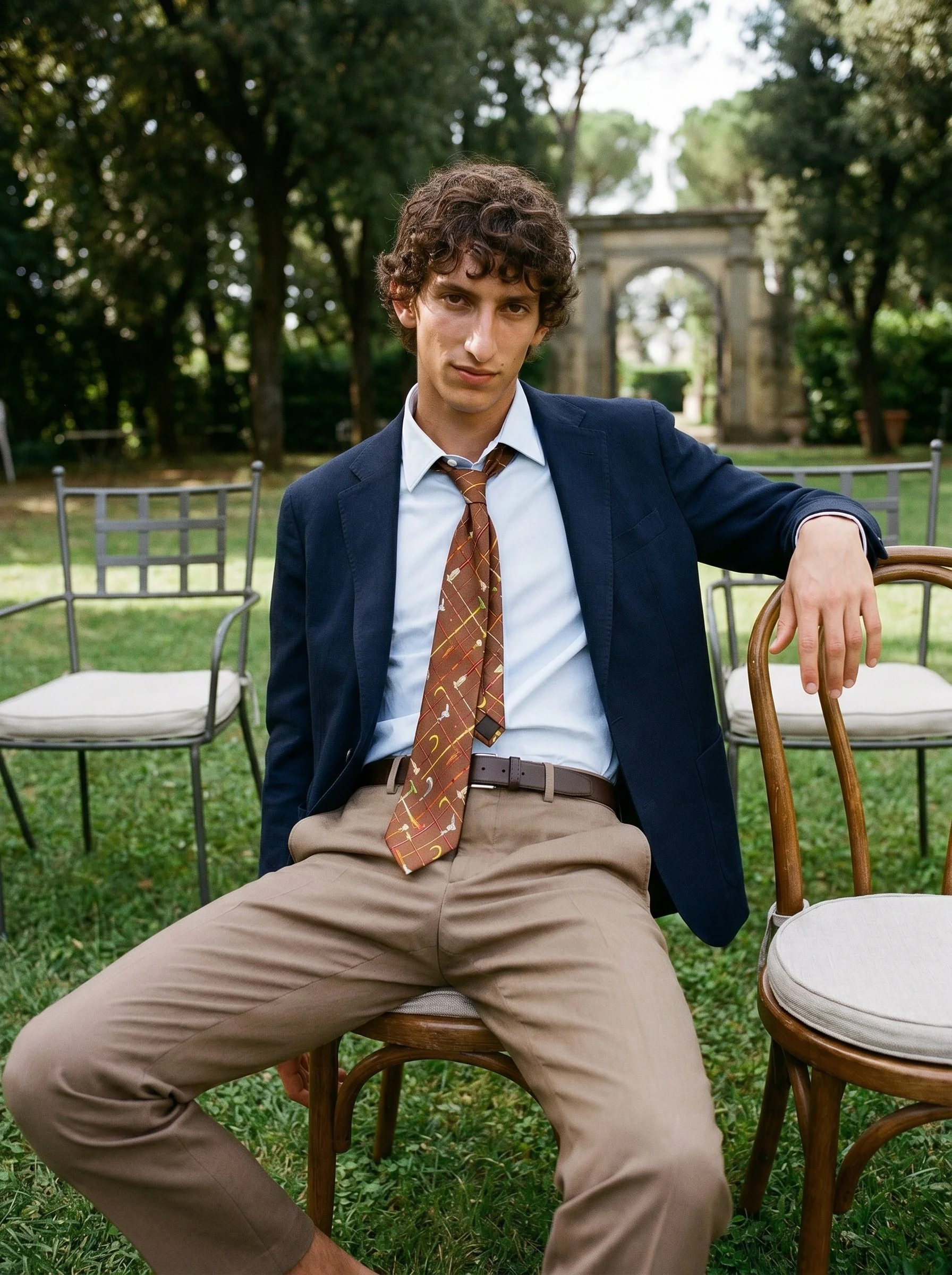 Young man with curly brown hair sitting outdoors at a garden party, wearing a navy blazer, white shirt, patterned tie, and khaki pants. He has a relaxed posture with one arm resting on a chair and is looking at the camera. There are chairs and greene
