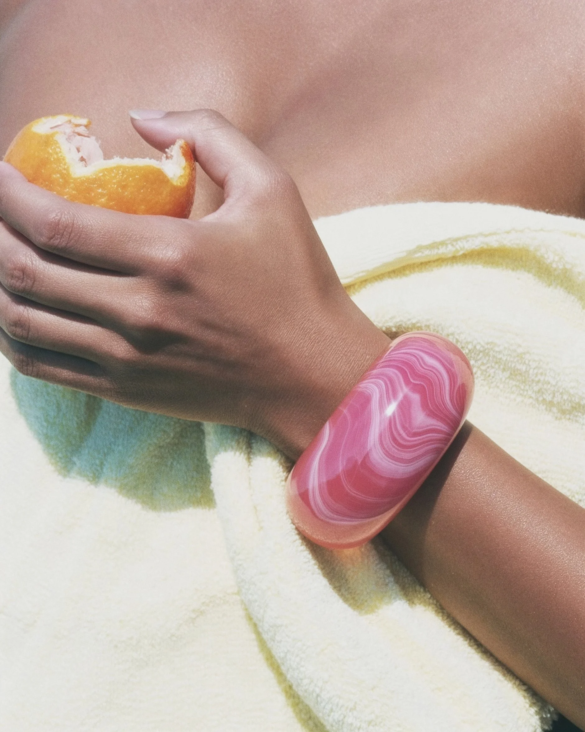 A person with brown skin peeling an orange with their fingers, wearing pink and white swirling patterned nail polish, resting on a yellow towel.  AI fashion editorial.