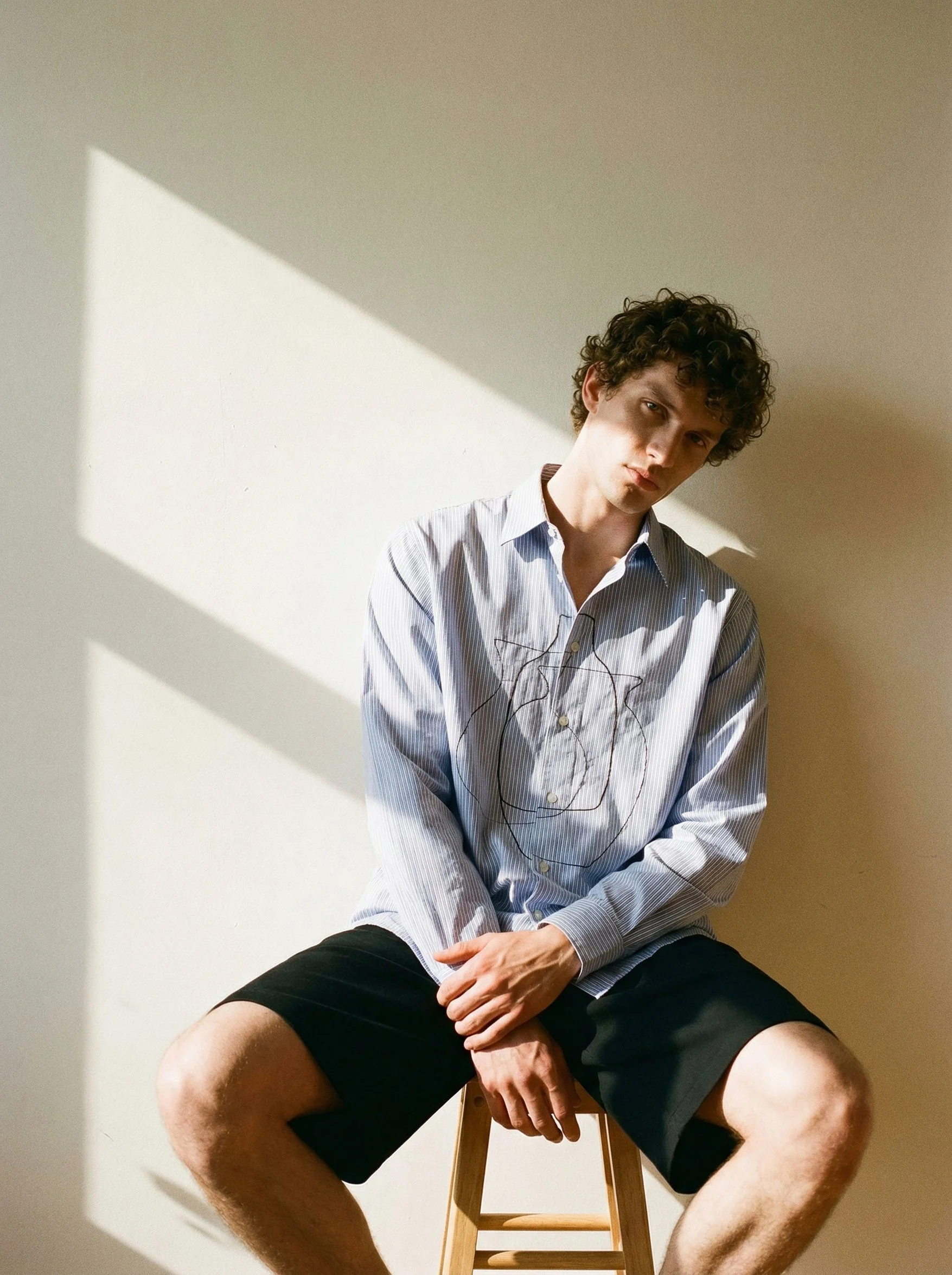 A young man with curly hair and light skin sits on a wooden stool against a plain, light-colored wall. He's wearing a blue and white striped button-up shirt and black shorts. The man has an intense, serious expression as he looks directly at the came