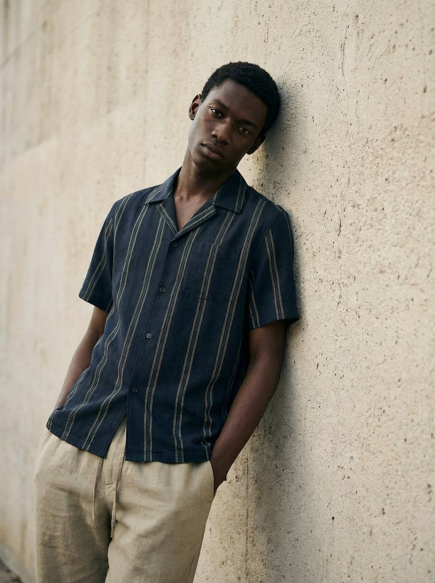 A young man with dark skin and short black hair leaning against a beige textured wall. He is wearing a dark blue striped short-sleeve button-up shirt and beige pants.