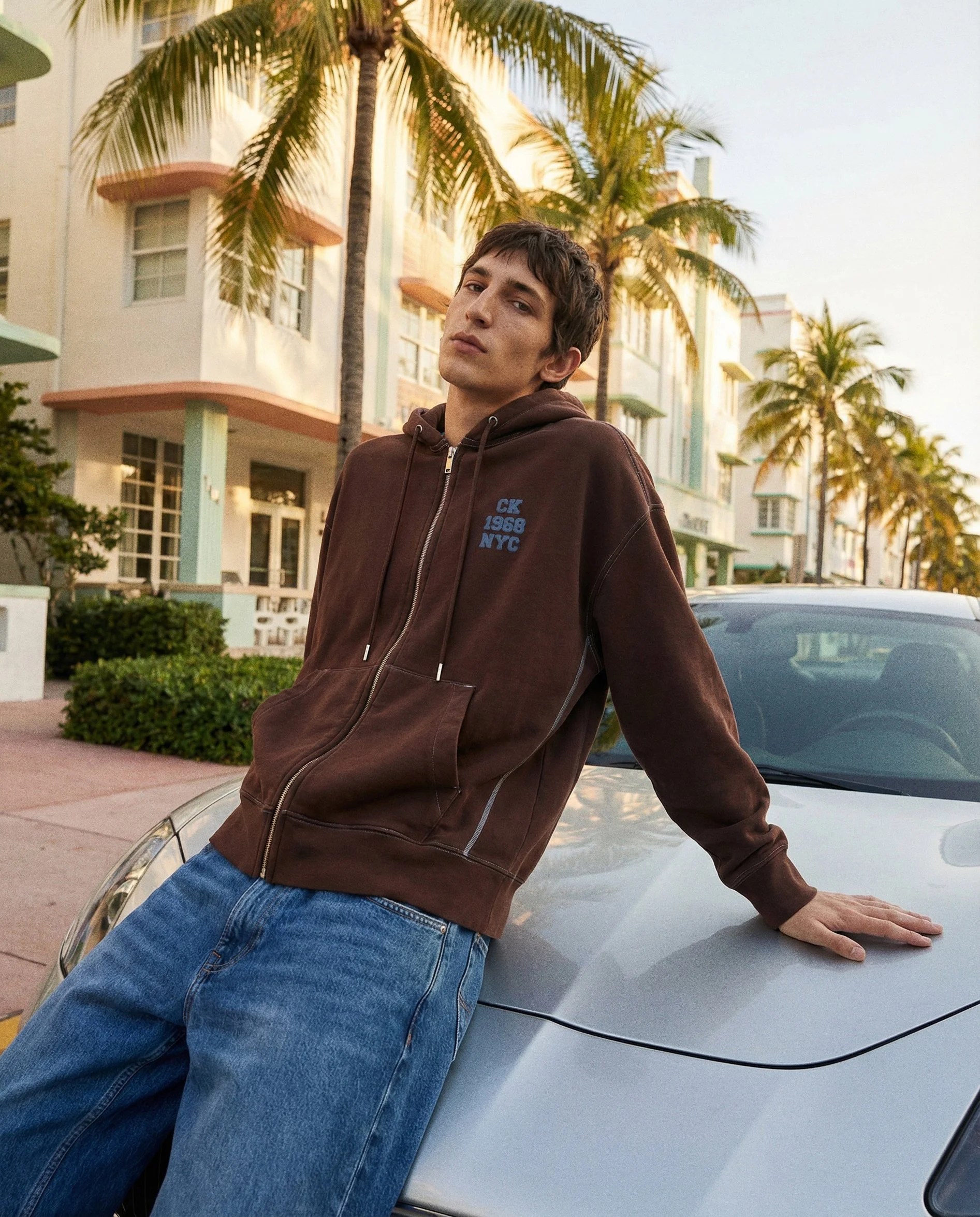 A young man with dark hair and light skin leaning against a silver car, wearing a brown hoodie with blue text, jeans, and looking at the camera in front of palm trees and a pastel-colored building during sunset.