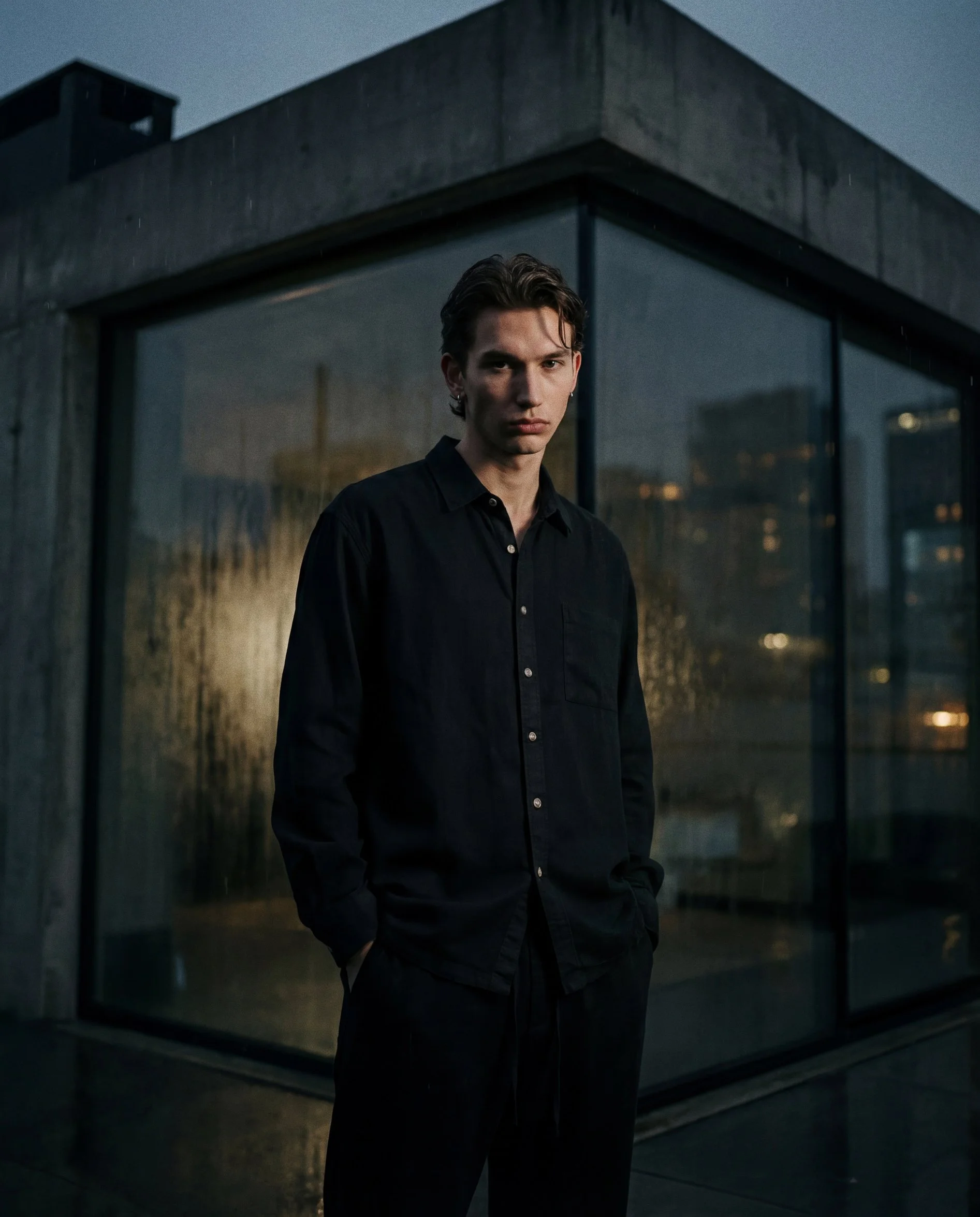 A young man in a black button-up shirt stands in front of a modern building with glass walls at dusk. The scene is slightly dark, with faint city lights visible in the background.